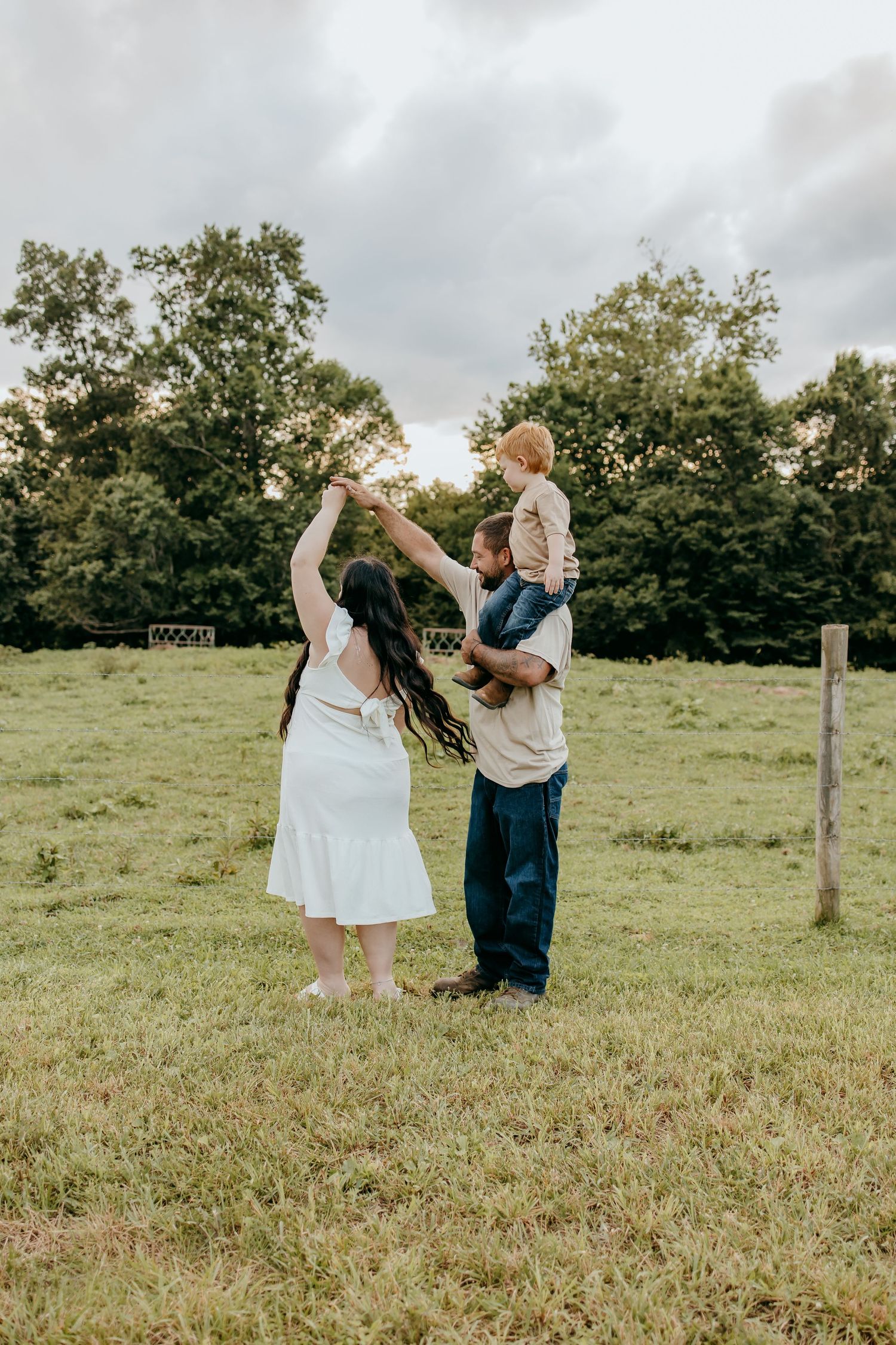 A family of three enjoys a playful moment in a green field, with a woman in a white dress, a man, and a child on his shoulders.