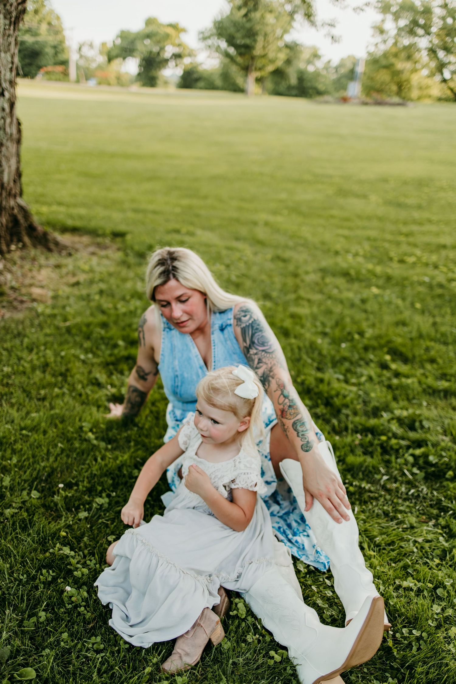 A tattooed blonde mother sits on green grass with her toddler daughter in a white dress at a sunny park.