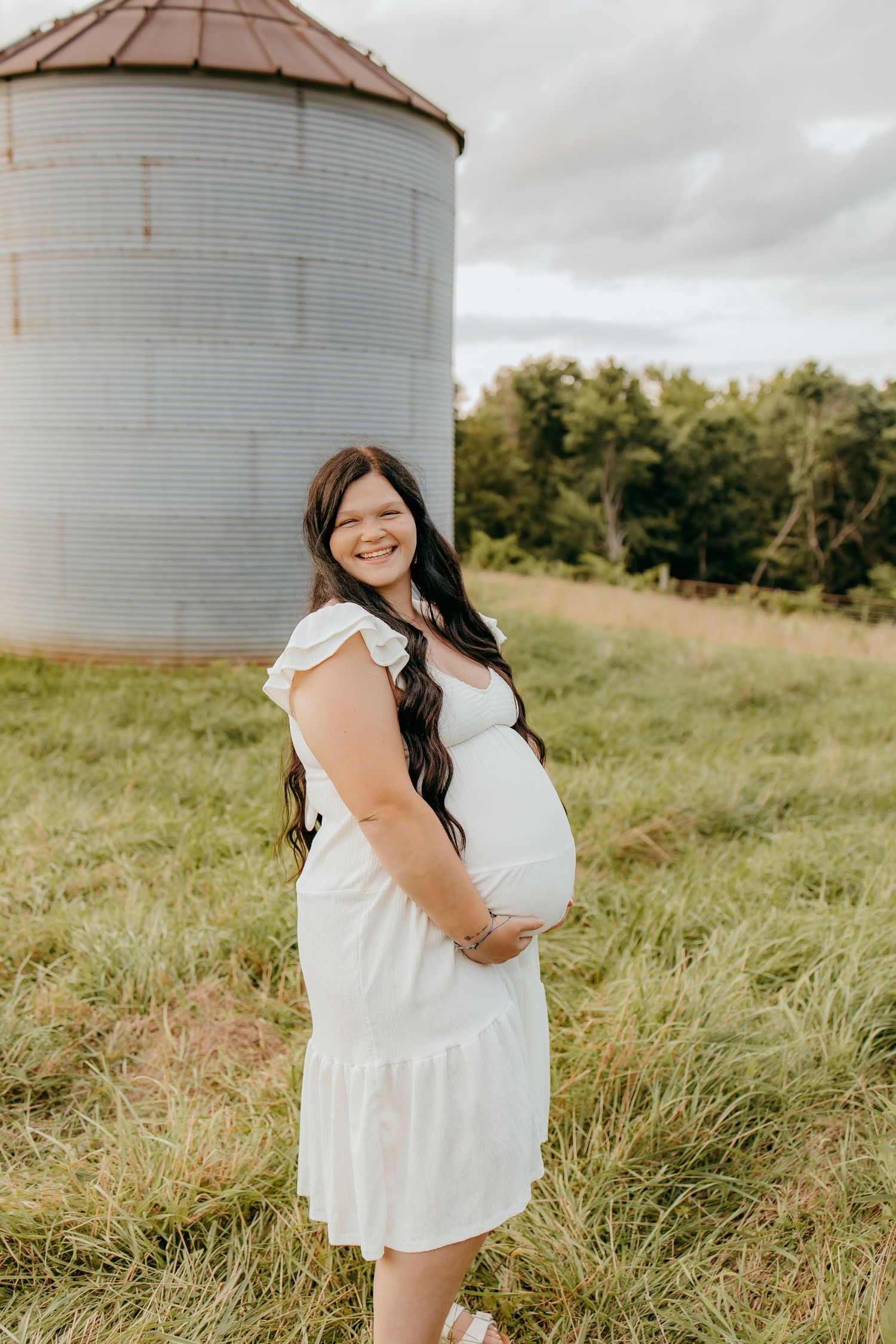 Smiling pregnant woman in white dress cradling baby bump outdoors near rustic grain silo on green farm field.