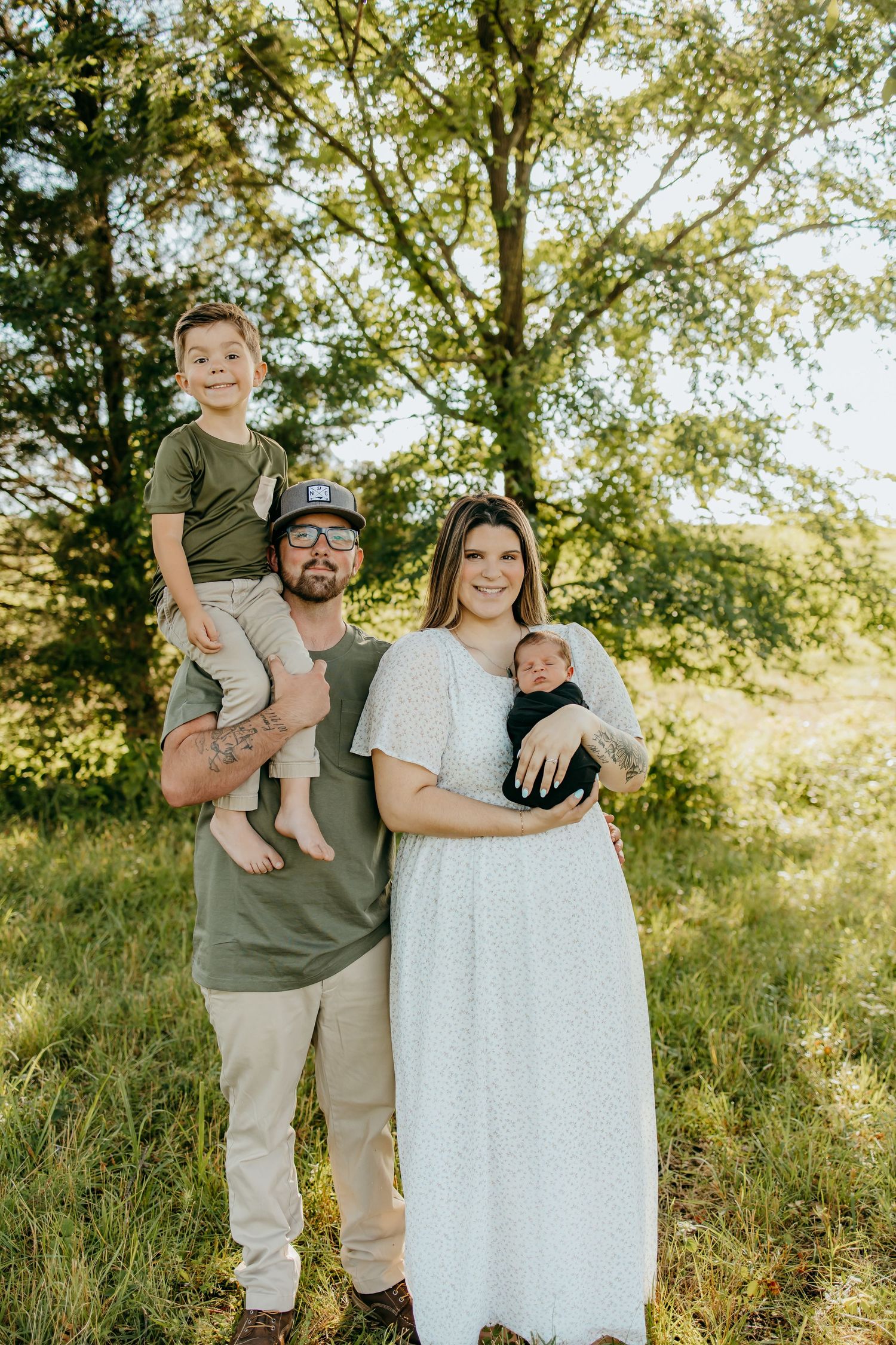 A young family of four poses outdoors, parents holding two young children, smiling in a lush green field.