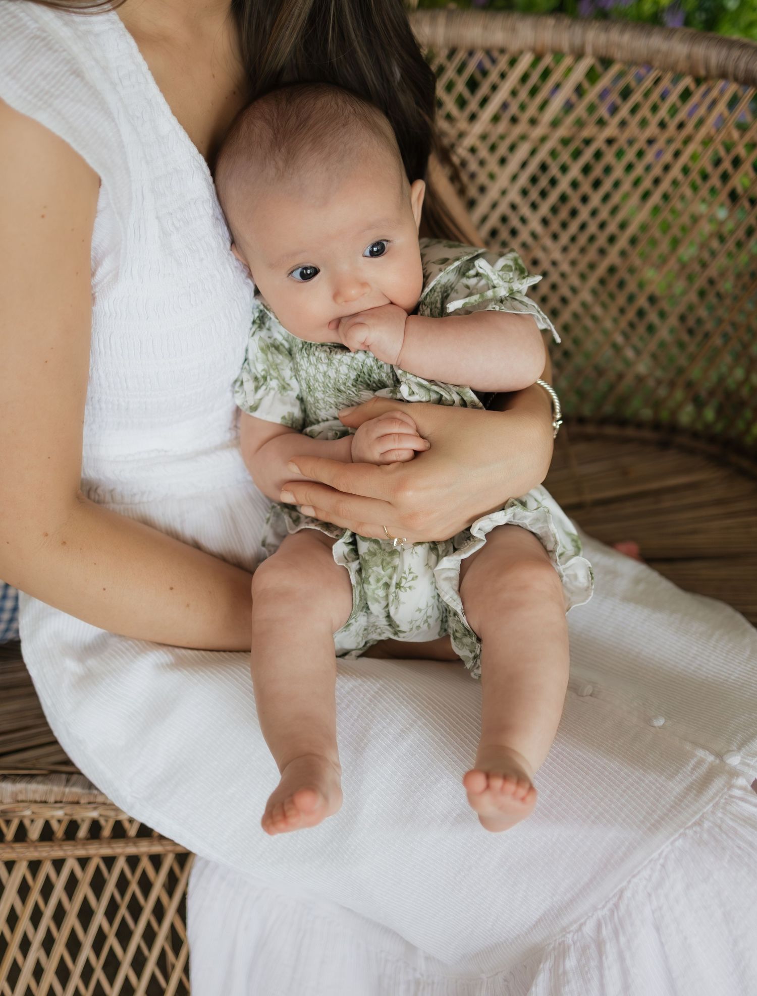 A mother in a white dress holds her baby dressed in a green floral romper while sitting in a wicker chair.
