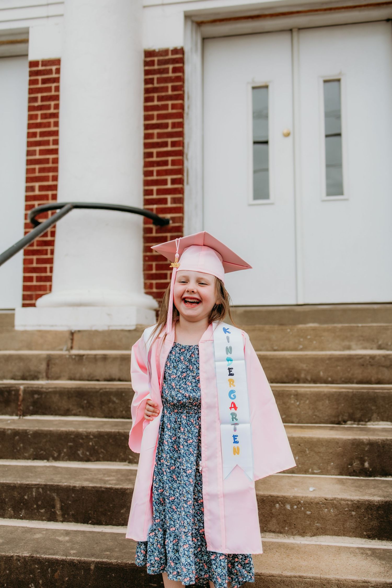 Happy girl in pink kindergarten graduation cap and gown laughing on steps outside brick building.