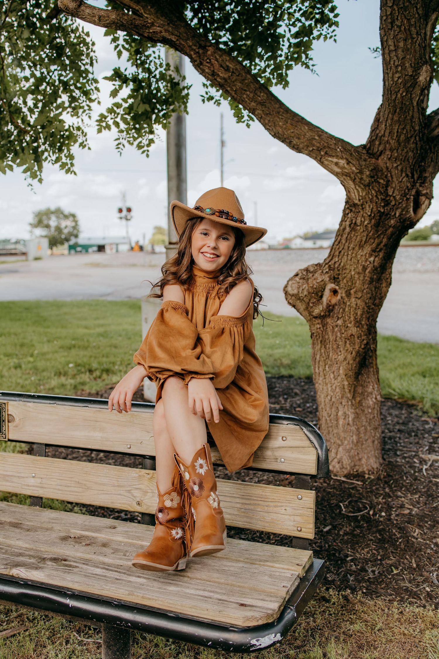 Young girl in brown dress, hat, and floral cowboy boots sits smiling on a wooden park bench beside a tree.