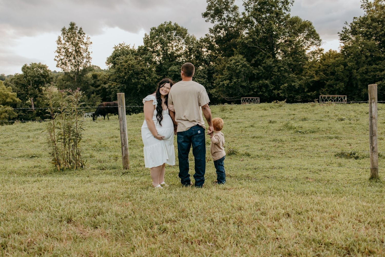 Family of three standing in a green pasture near a wooden fence, surrounded by lush trees on a cloudy day.