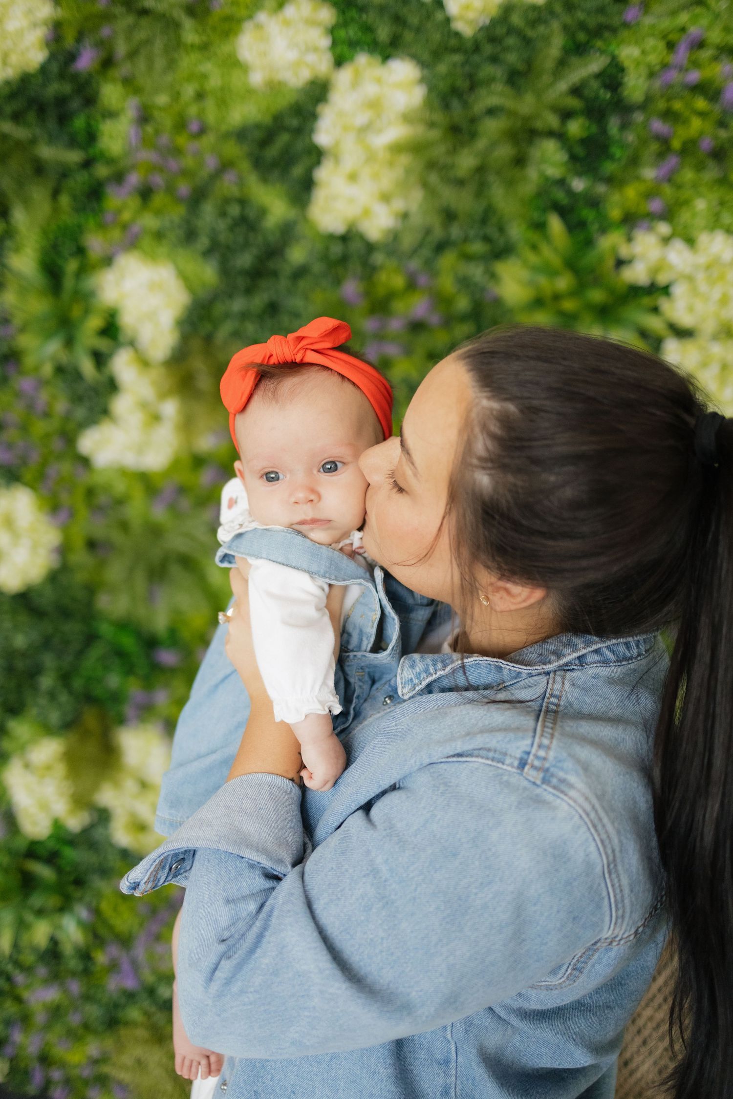 A young mother in a denim jacket kisses her baby girl wearing a red bow headband against a floral green wall.