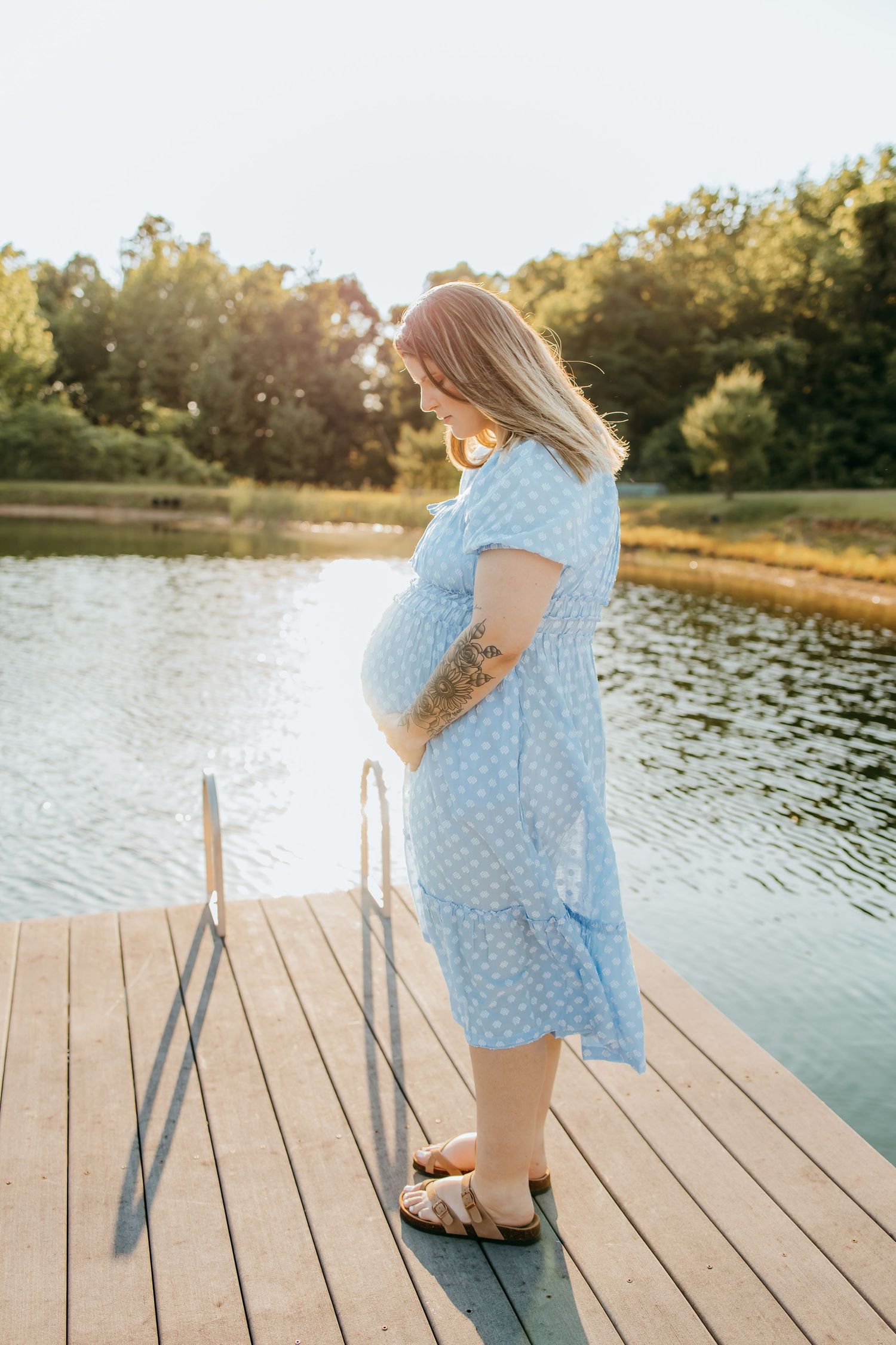 Pregnant woman in blue polka dot dress standing on wooden dock by lake, cradling baby bump in golden sunlight.