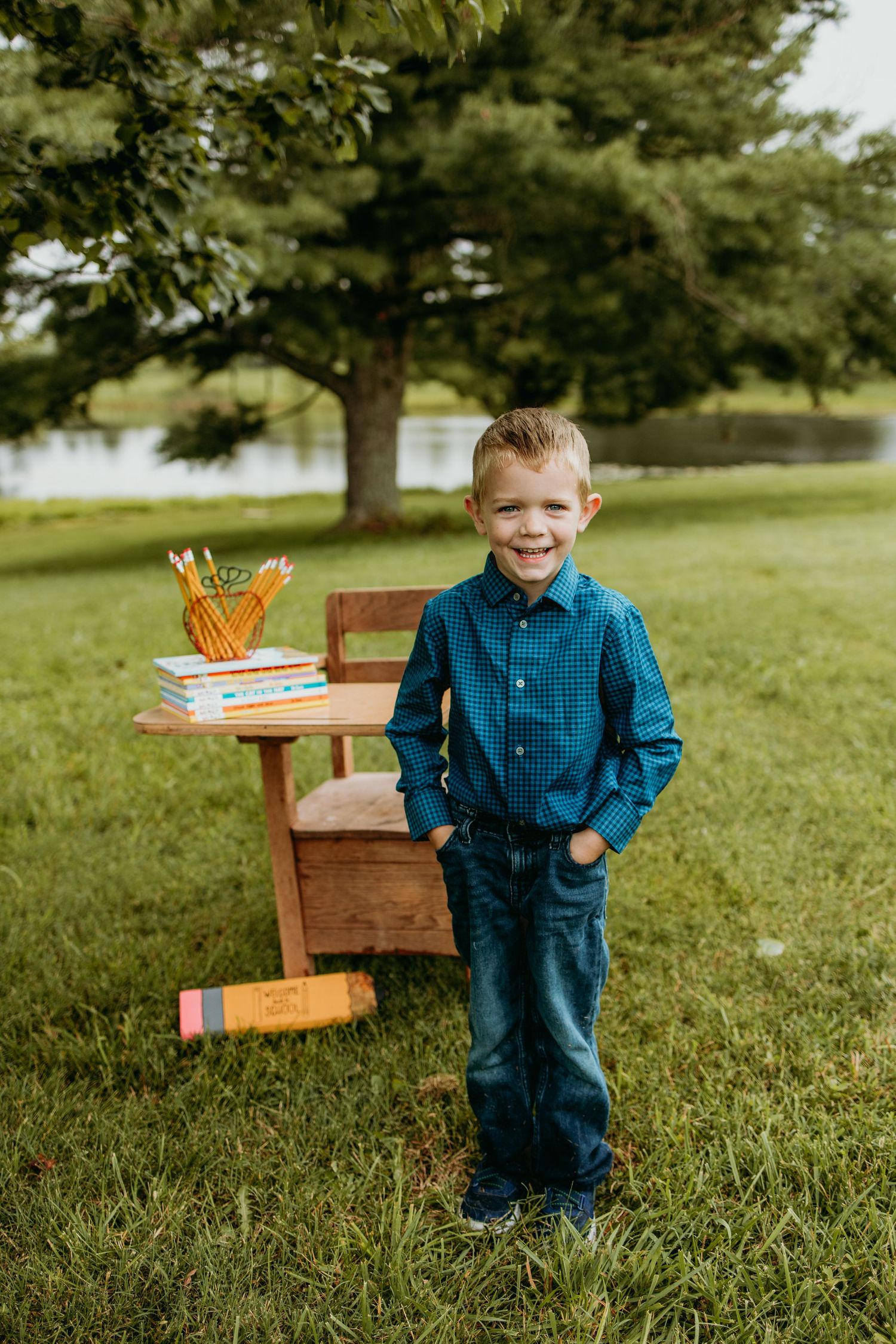 Smiling young boy in blue plaid shirt and jeans standing beside a wooden school desk outdoors under a large tree.