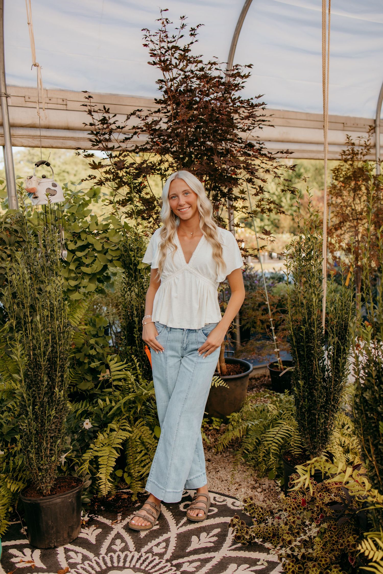 senior in white peplum top and light jeans poses among lush green plants in a sunlit greenhouse.