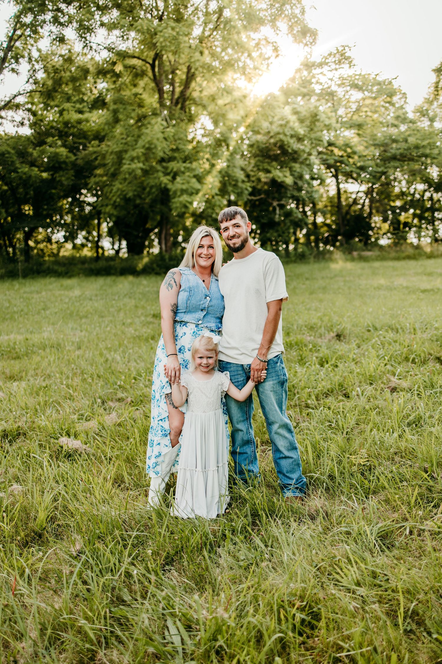 Family of three posing in a sunny green meadow, parents smiling with young daughter in white dress.