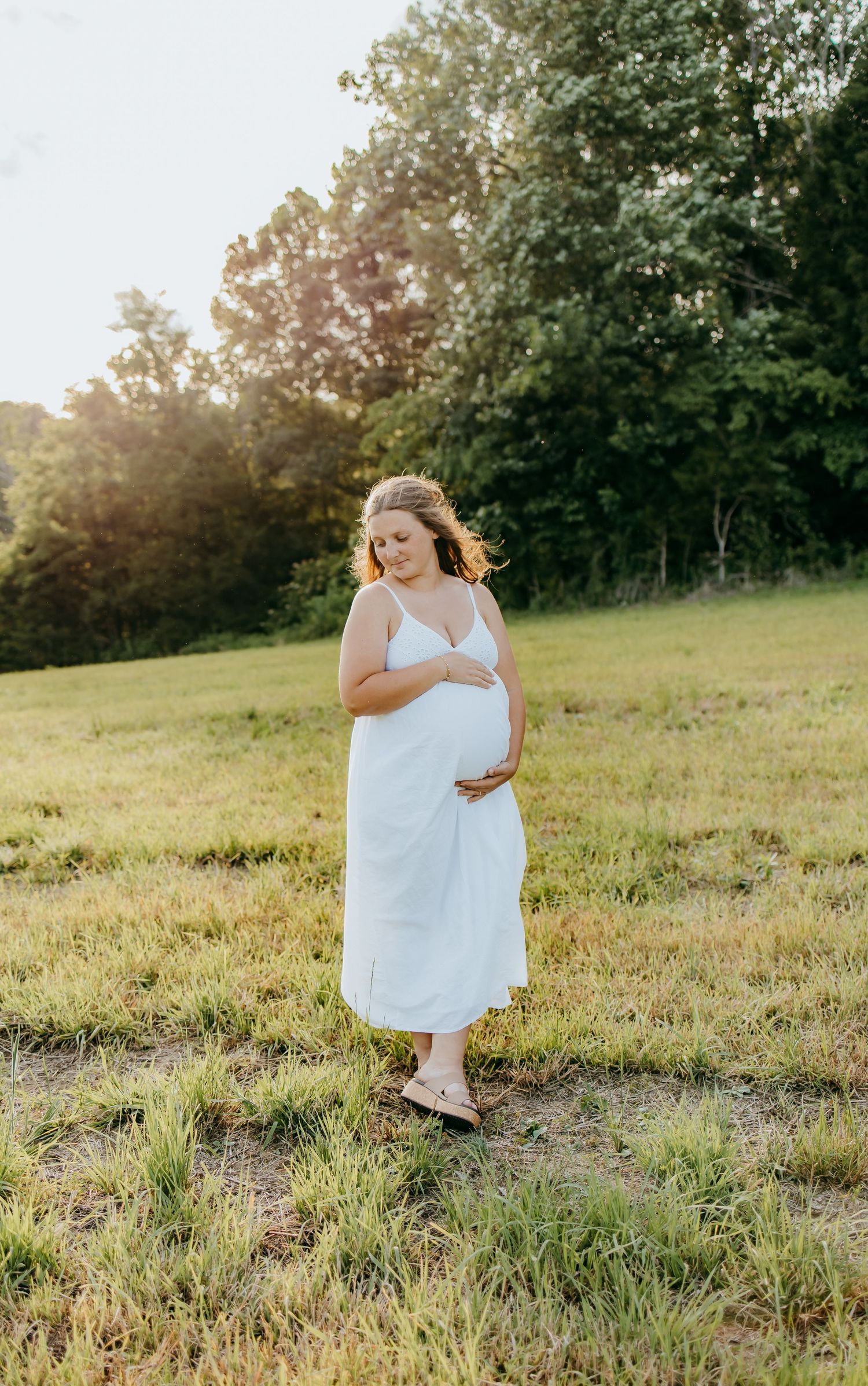 Pregnant woman in white dress cradling baby bump in sunlit green field with trees in background.