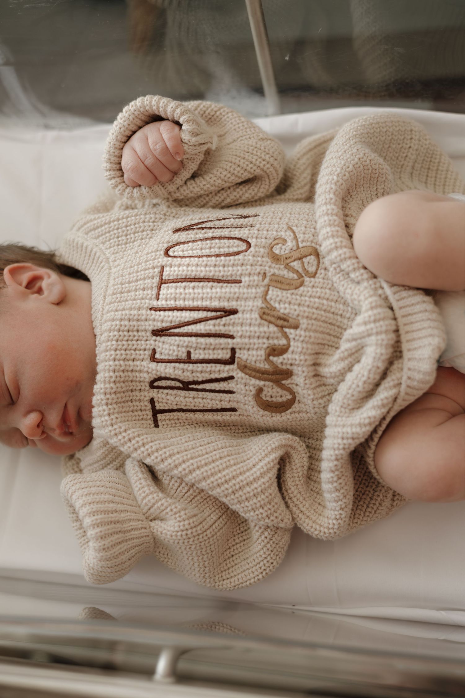 Sleeping newborn baby wearing a personalized beige knit romper embroidered with the name Trenton Chris.
