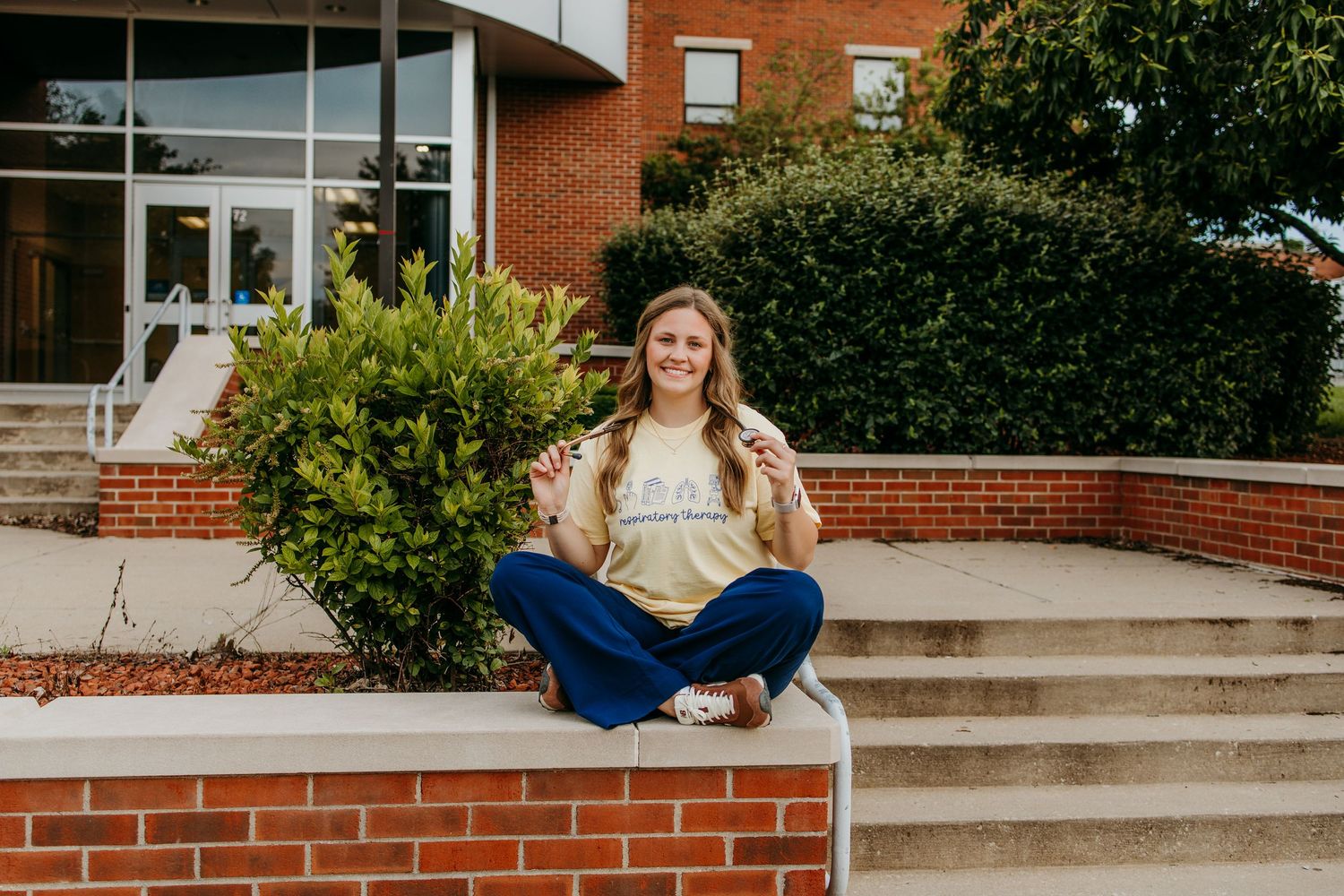 Smiling young woman sitting cross-legged on brick steps outside a school building, wearing a gold sweatshirt and jeans.