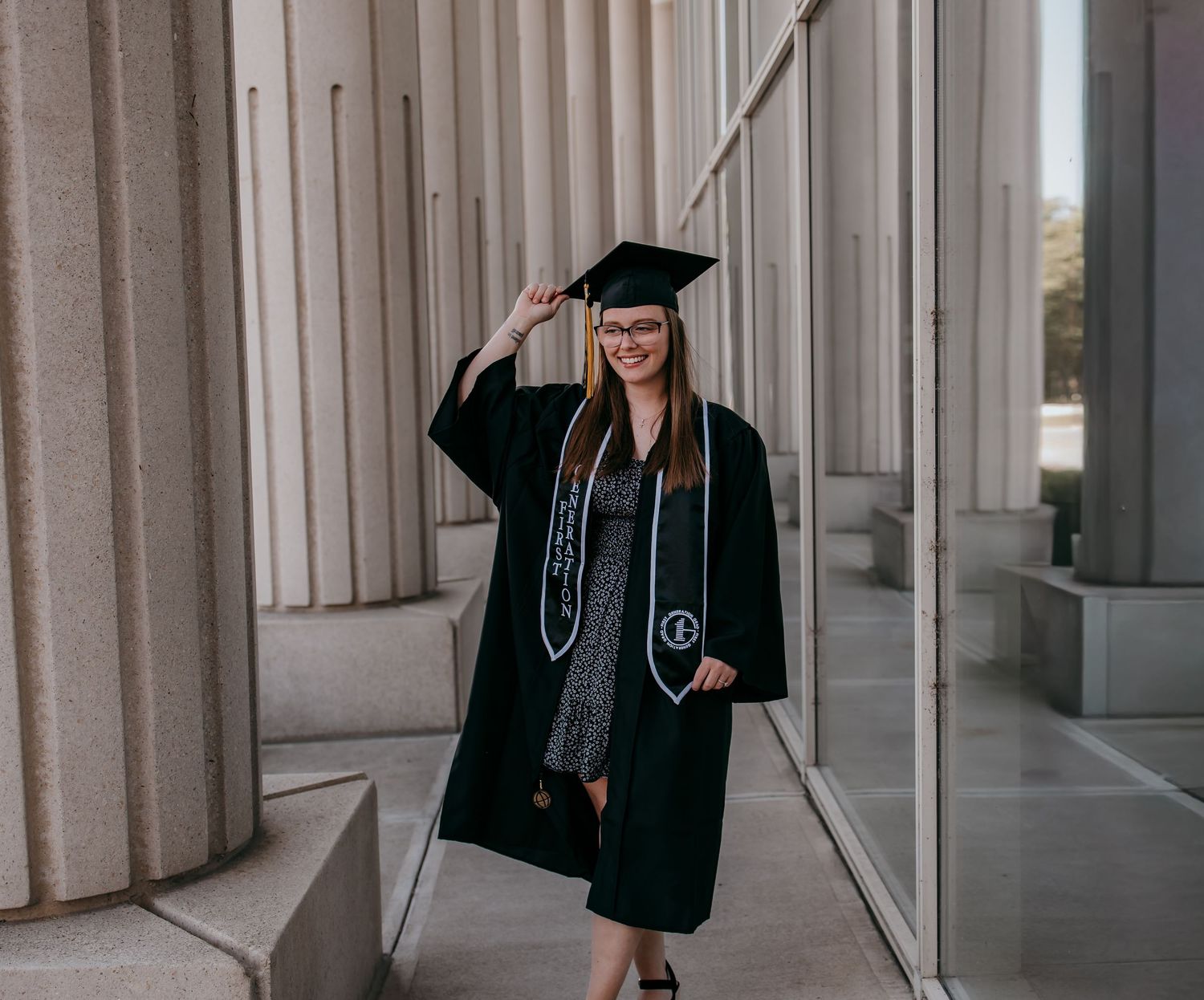 A smiling woman in black graduation cap and gown poses between large columns outdoors on a sunny day.