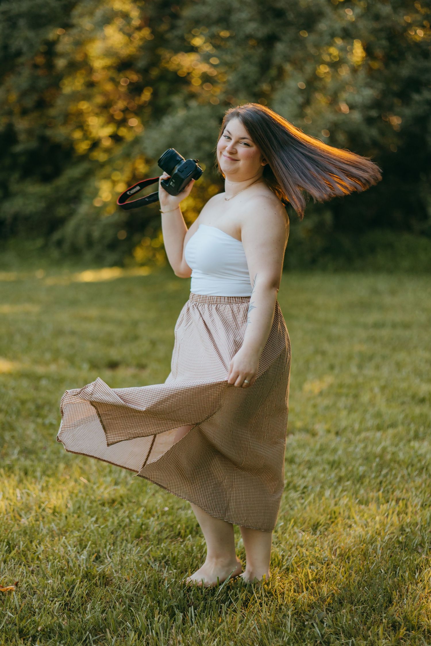 Plus-size woman in white tube top and beige maxi skirt twirling outdoors, holding a camera in a sunny park.