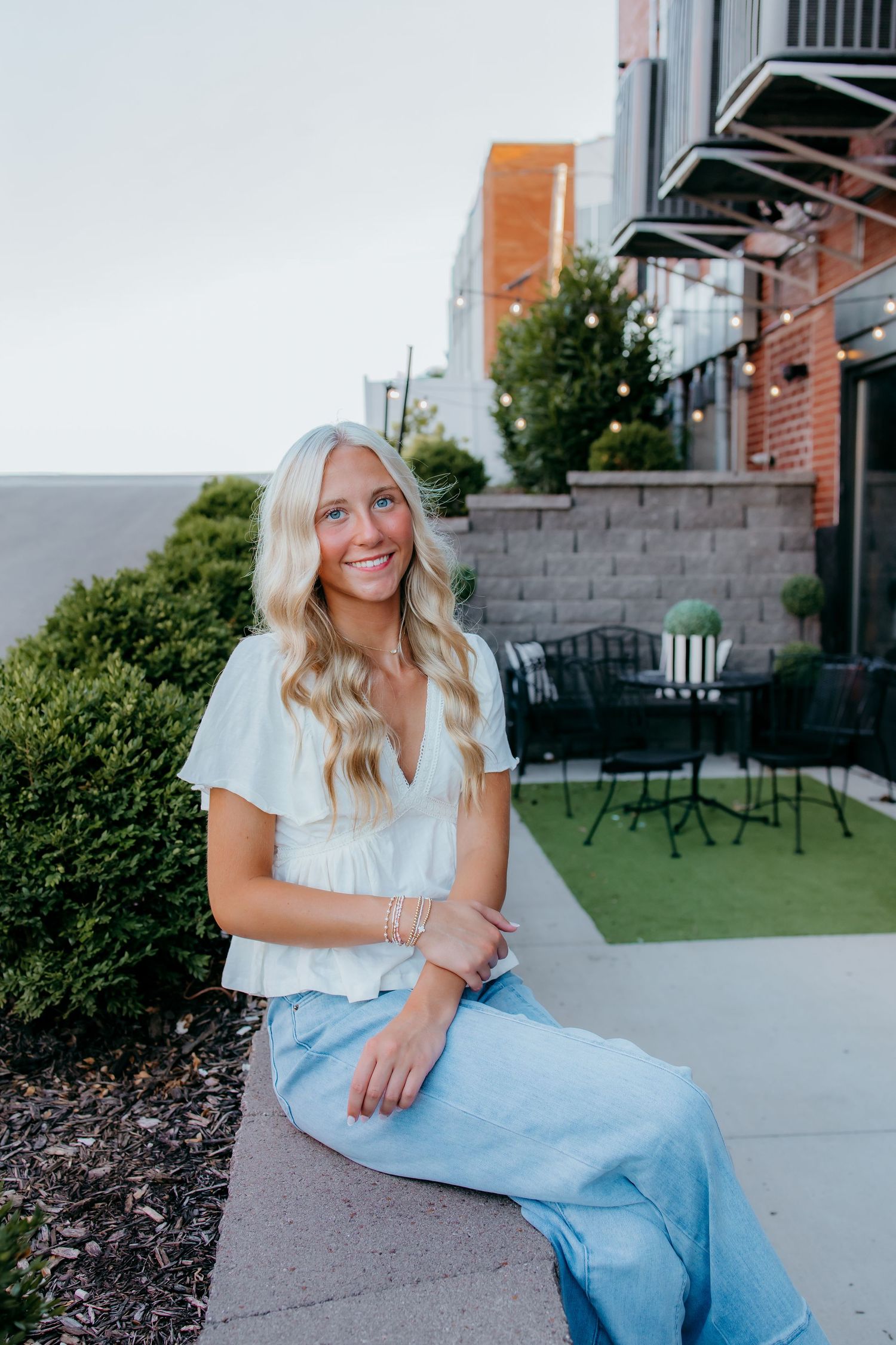 Senior smiling in white top and jeans, seated outdoors near a patio with greenery and string lights.