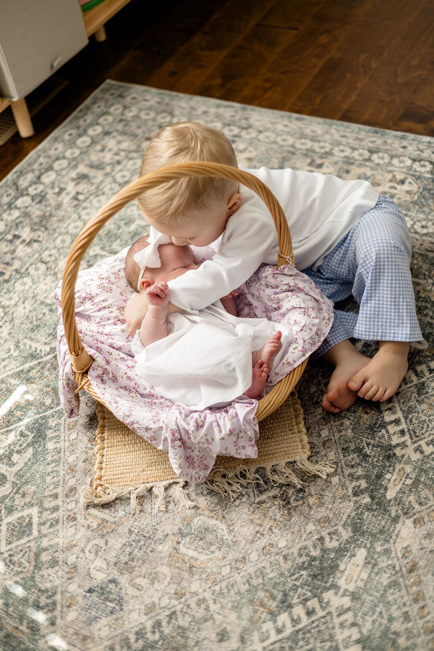 Toddler gently kissing newborn baby sibling in a wicker basket on a patterned rug.