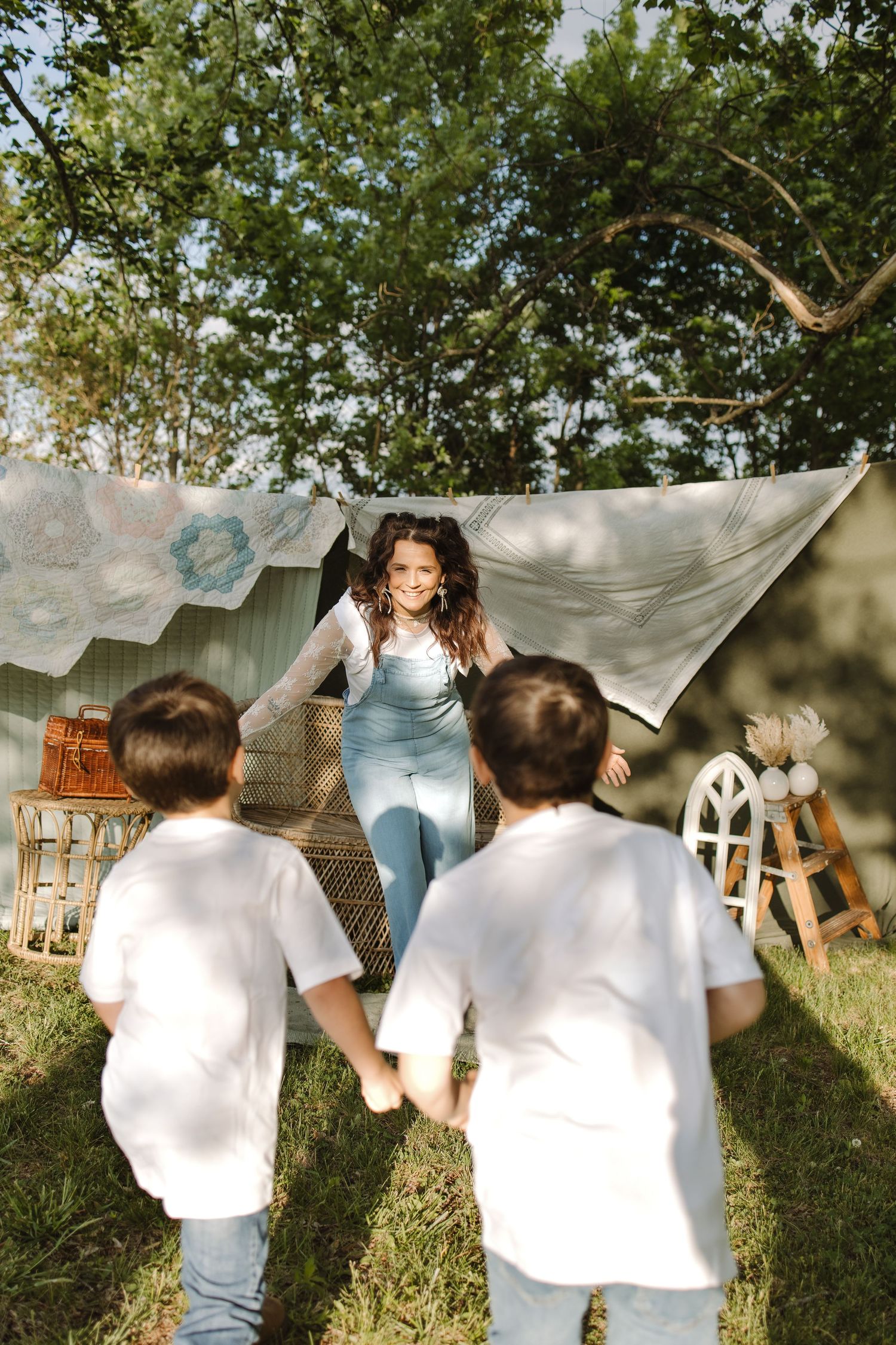 A smiling woman in denim overalls plays with two young boys in white shirts in a sunny backyard with draped fabric backdrop.