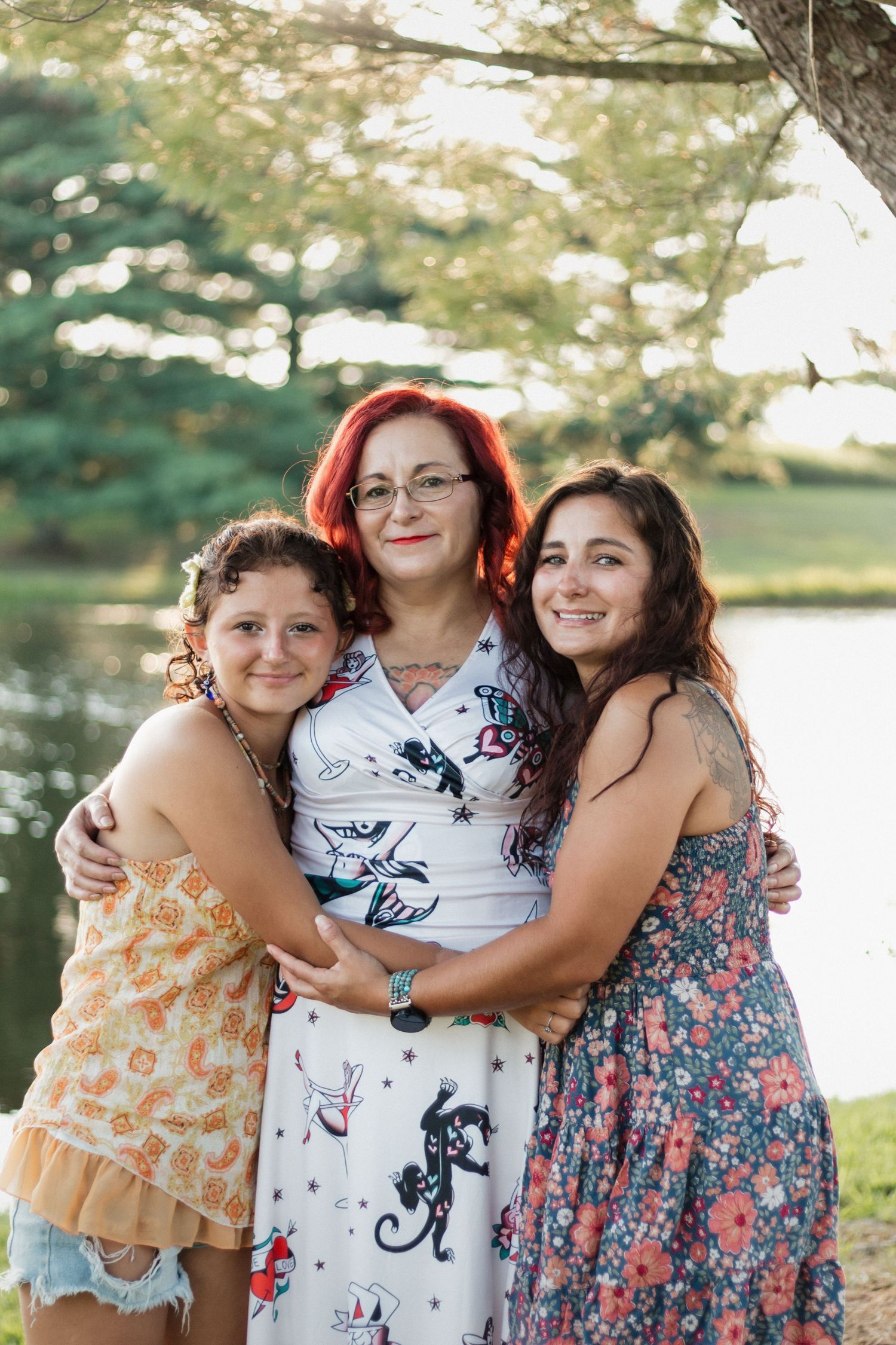A woman with red hair and two young girls smile and hug outdoors near a lake on a sunny day.