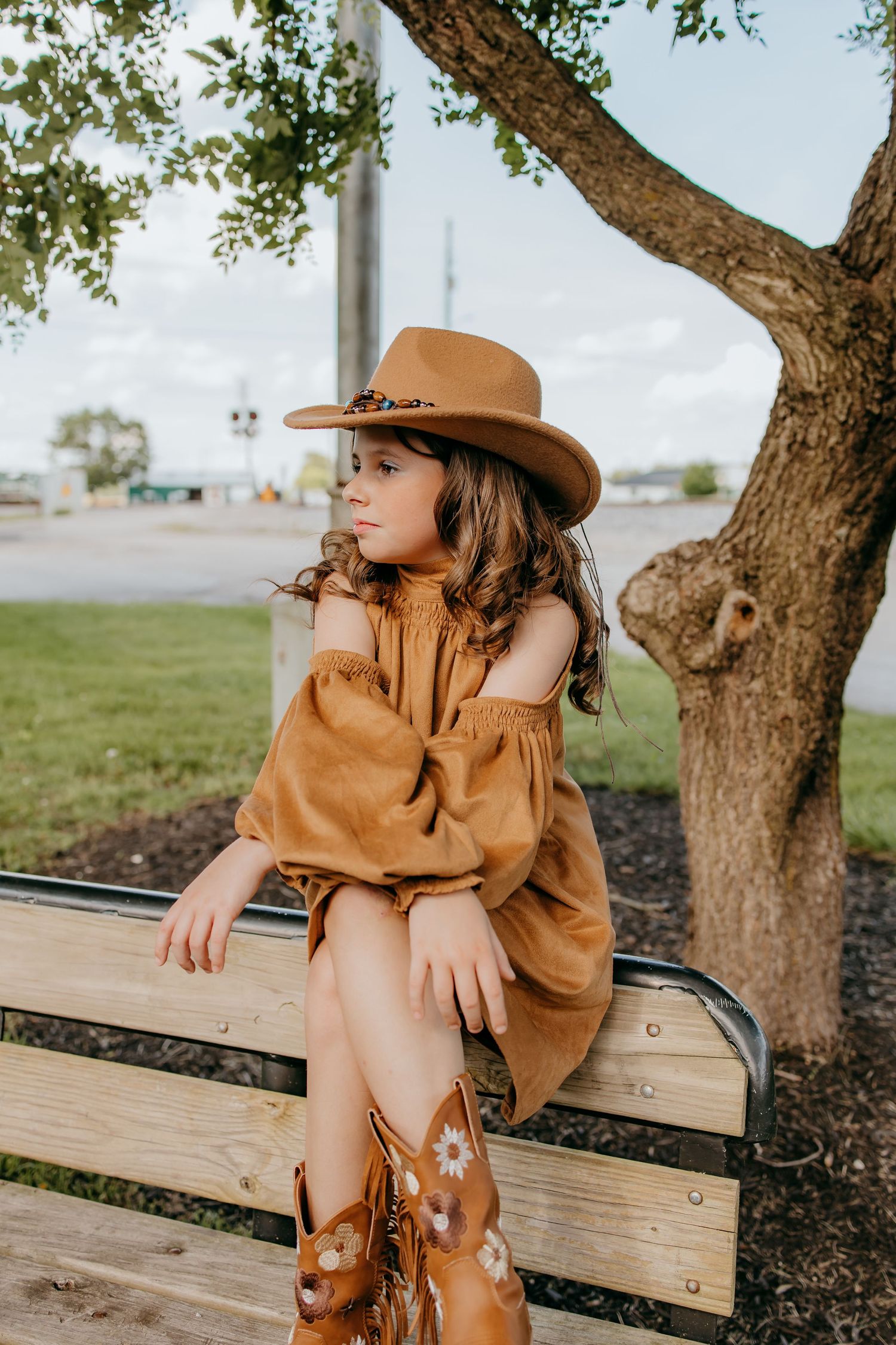 Young girl in brown cowboy hat, rust off-shoulder dress, and floral boots sits on bench under tree.