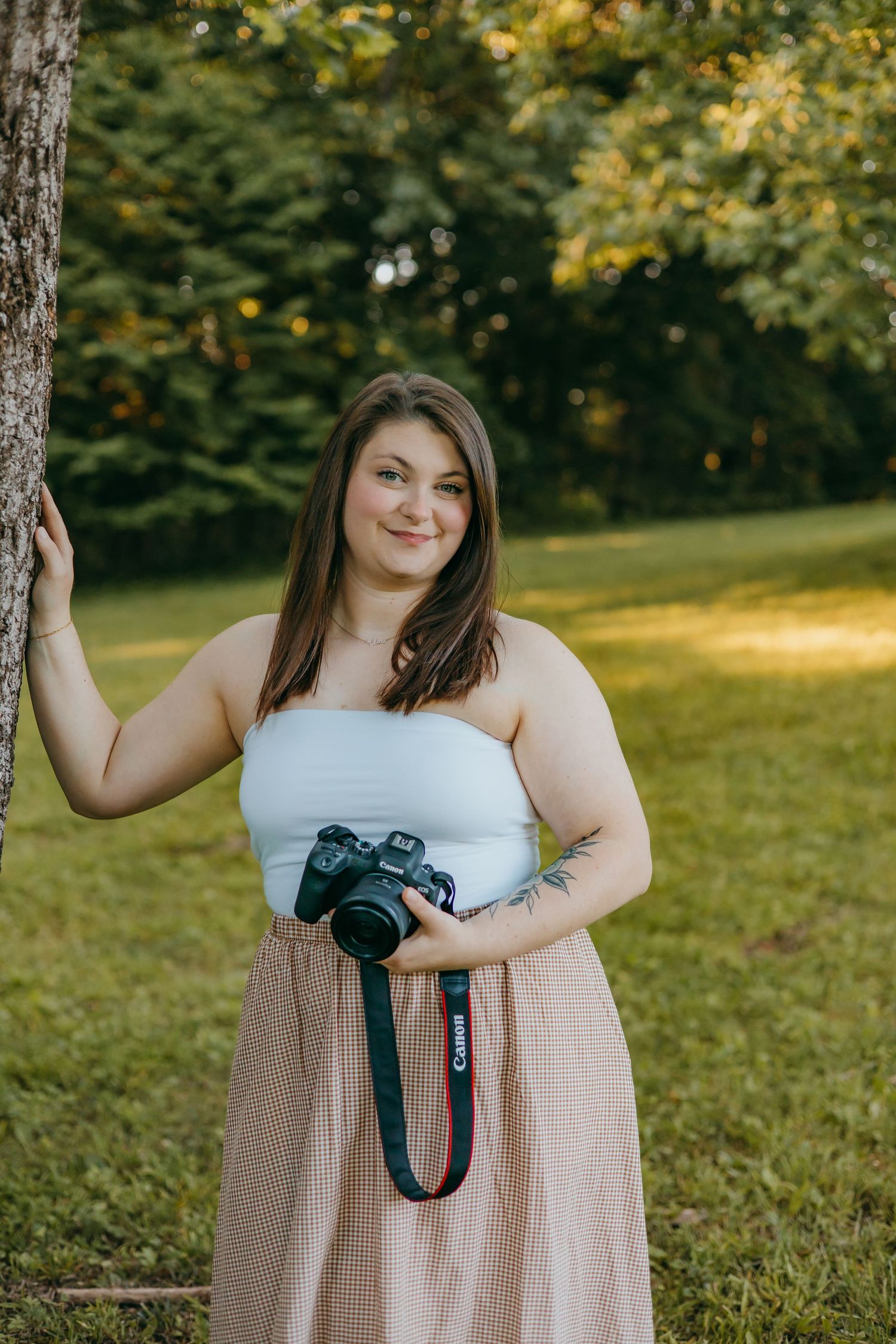 Woman in white tube top and plaid skirt holding a Canon camera, smiling outdoors in a sunlit green park.
