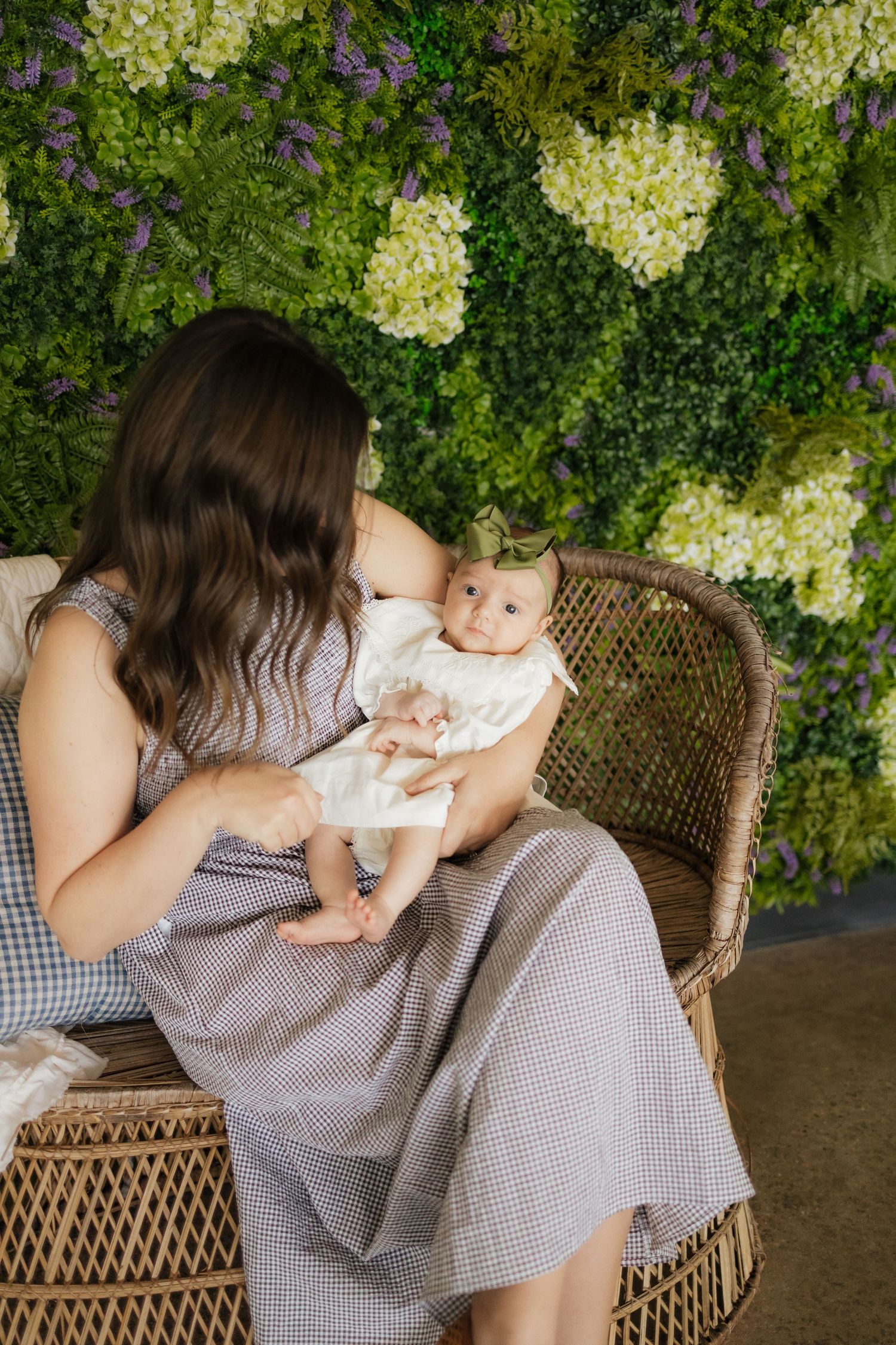 Mother in gray dress holds newborn baby in white outfit while sitting in wicker chair against floral green wall backdrop.