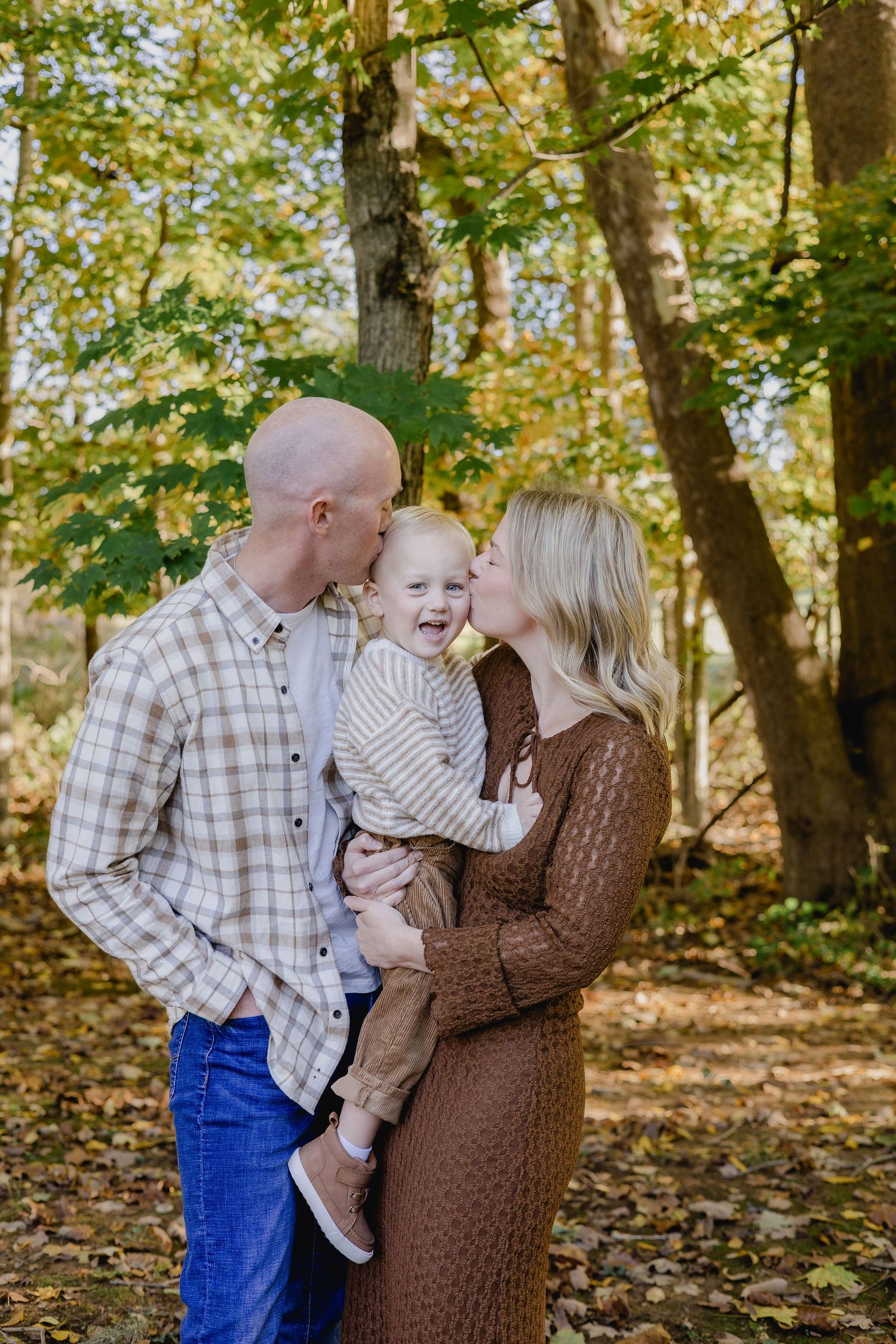 A happy family of three poses outdoors in autumn woods, parents kissing their laughing toddler on the cheeks.