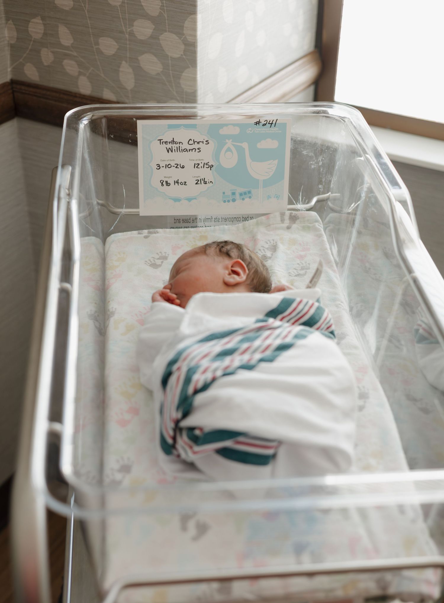 Newborn baby swaddled in striped blanket sleeping peacefully in a hospital bassinet with name card attached.