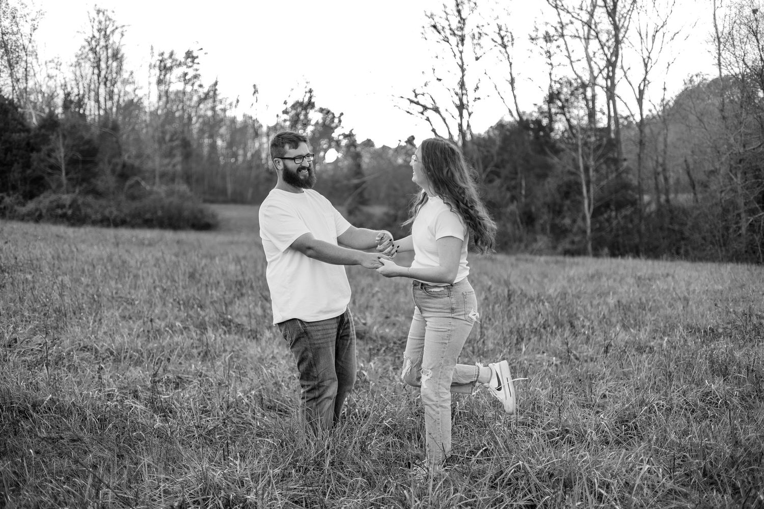 Black and white photo of a couple holding hands and dancing together in a grassy field with trees in the background.
