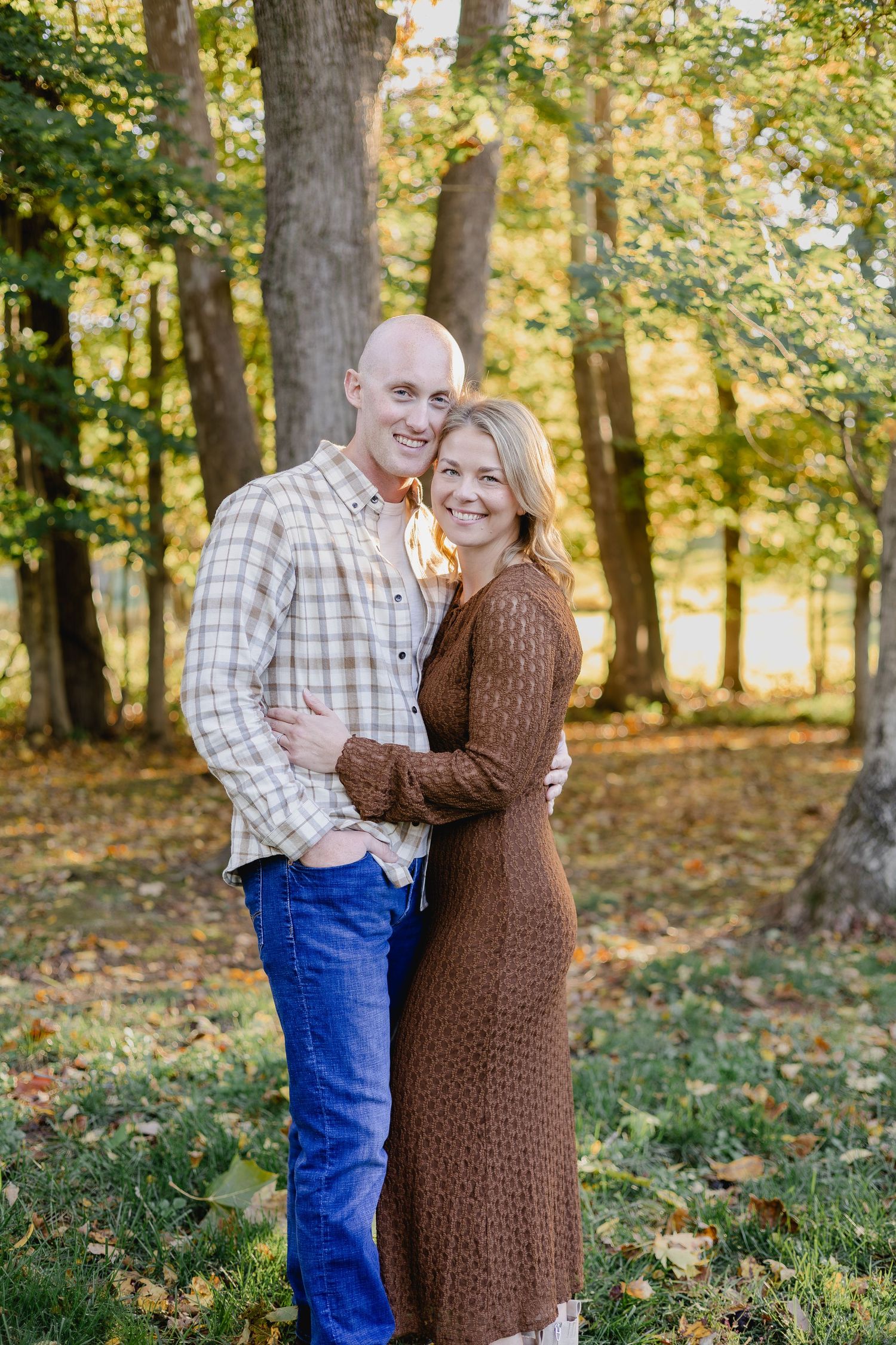 A smiling couple embraces outdoors in a sunlit forest during autumn, wearing casual fall attire.