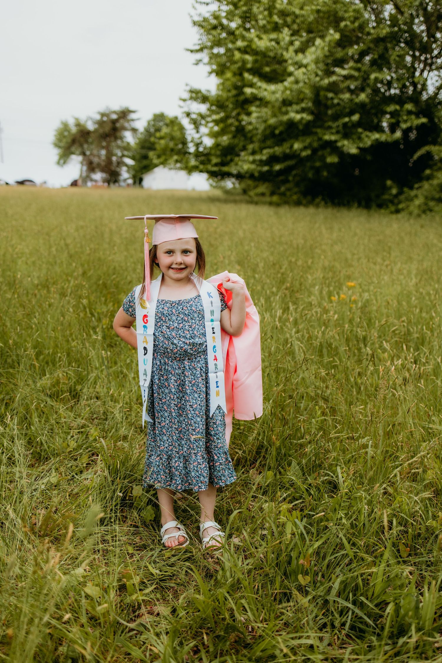Young girl in graduation cap, sash, and floral dress stands smiling in green field, holding pink diploma.