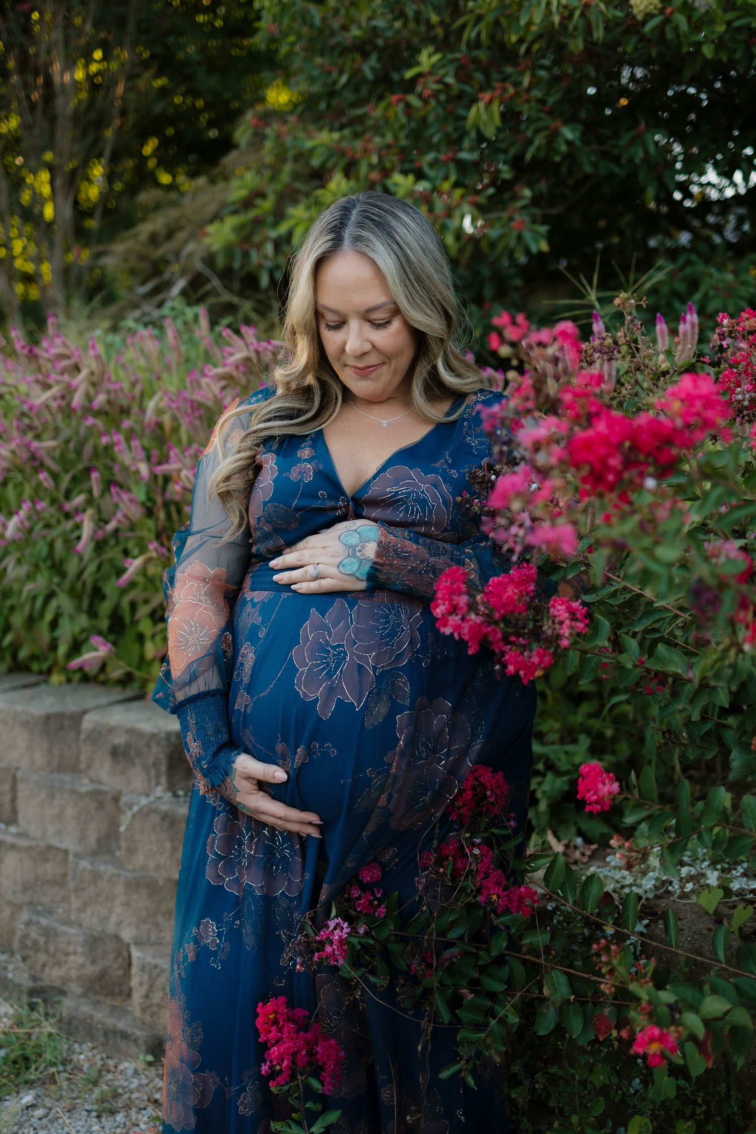 Pregnant woman in blue floral maxi dress cradling baby bump surrounded by pink blooming flowers in a garden.