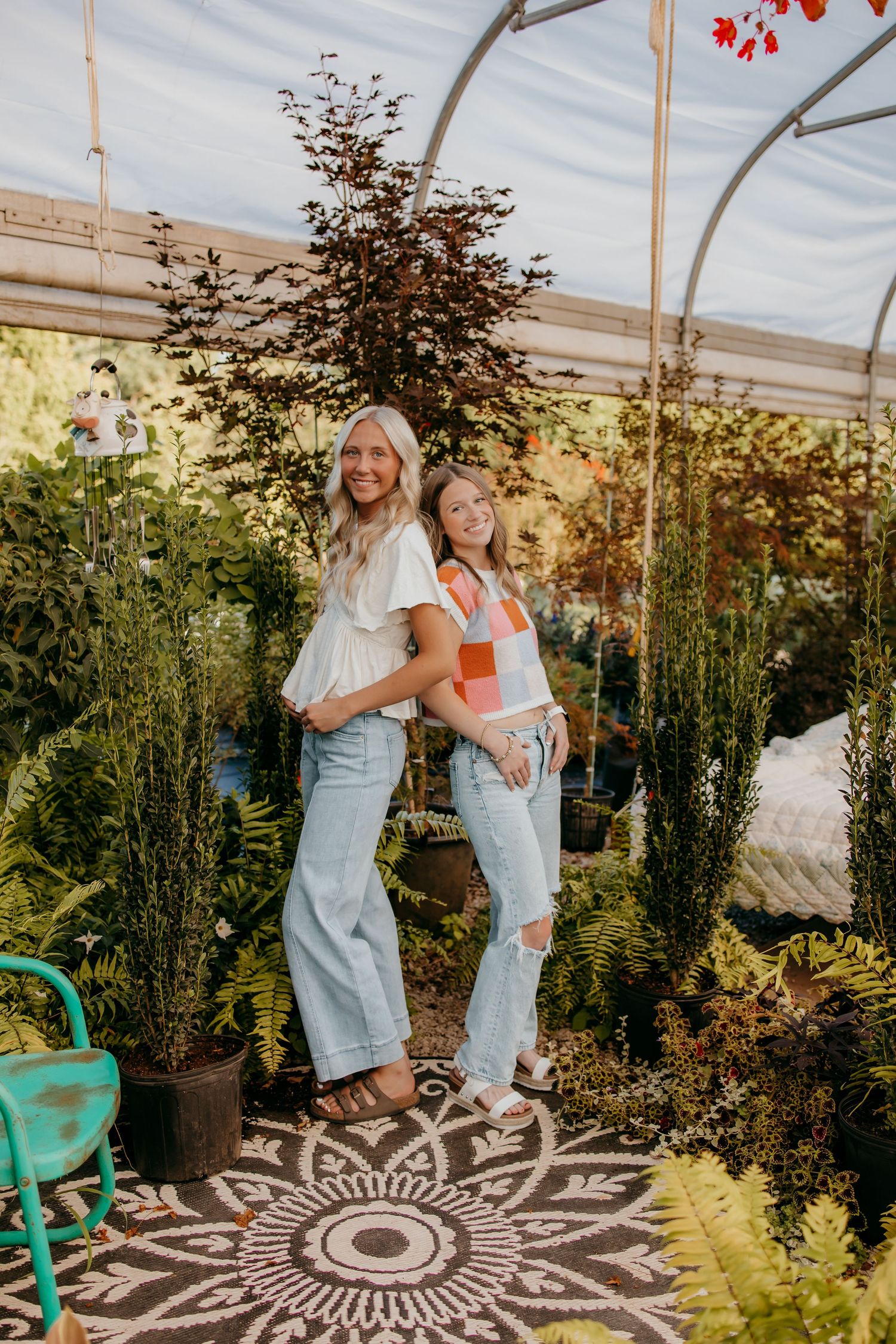 Two smiling seniors in jeans pose together inside a lush greenhouse nursery on a decorative patterned rug.