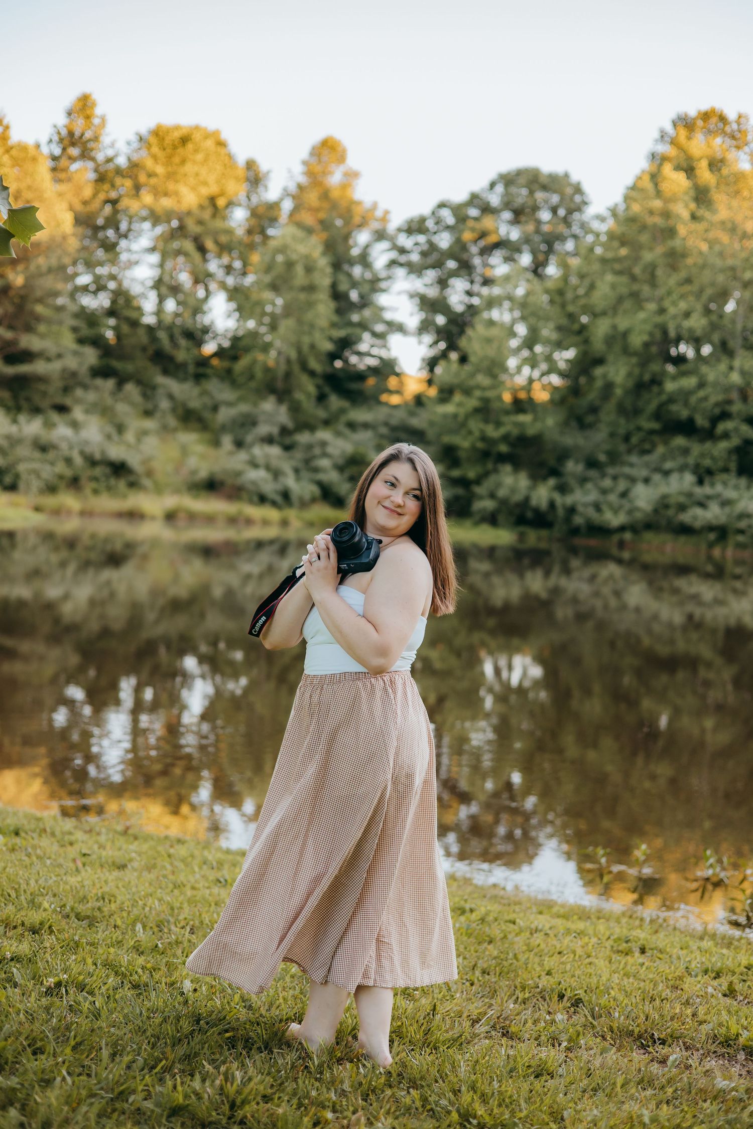 Young woman in plaid maxi skirt and white top poses by a reflective pond surrounded by lush green trees.
