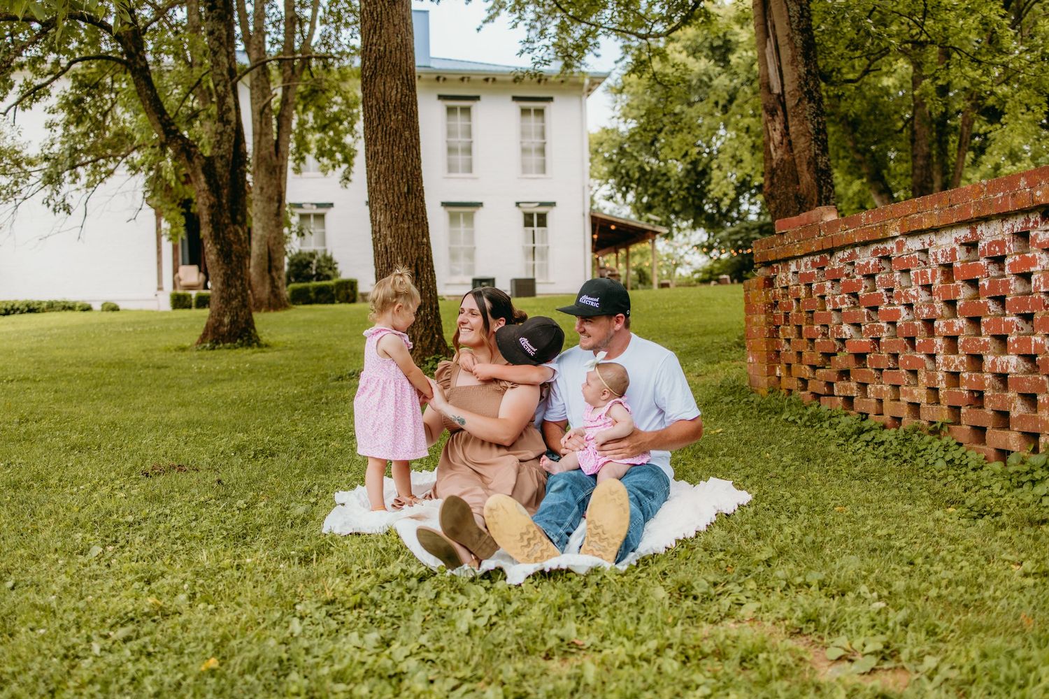 Family of five sitting on a white blanket in a lush green yard near trees and a historic white building.