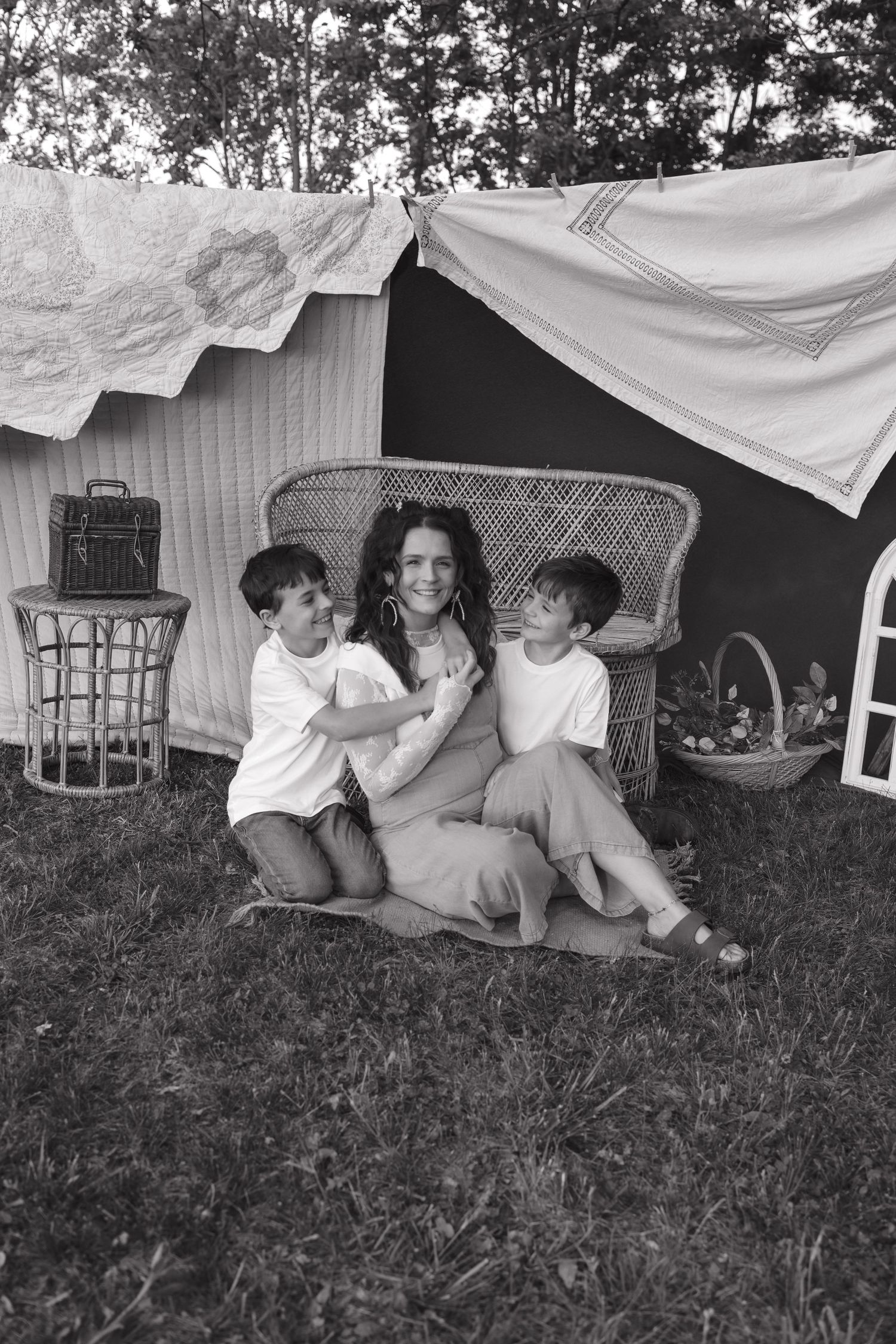 Black and white photo of a smiling woman sitting on grass with two young boys beside a makeshift blanket fort outdoors.