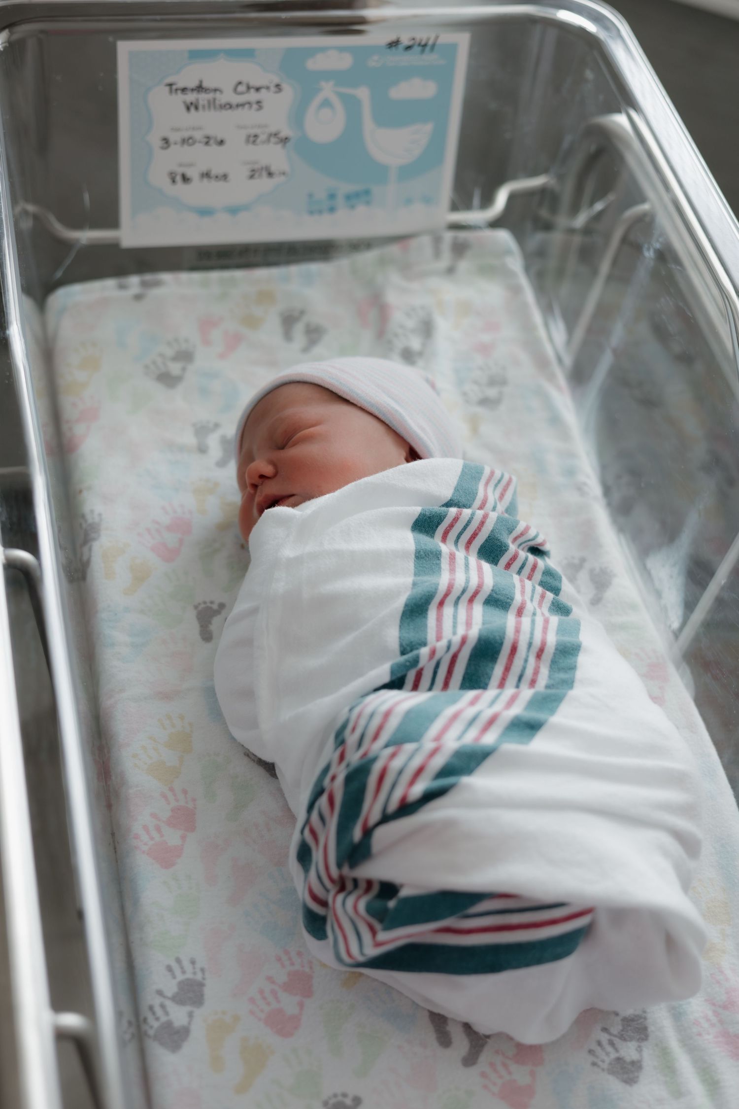 Newborn baby swaddled in striped hospital blanket sleeping peacefully in a clear bassinet with name card.
