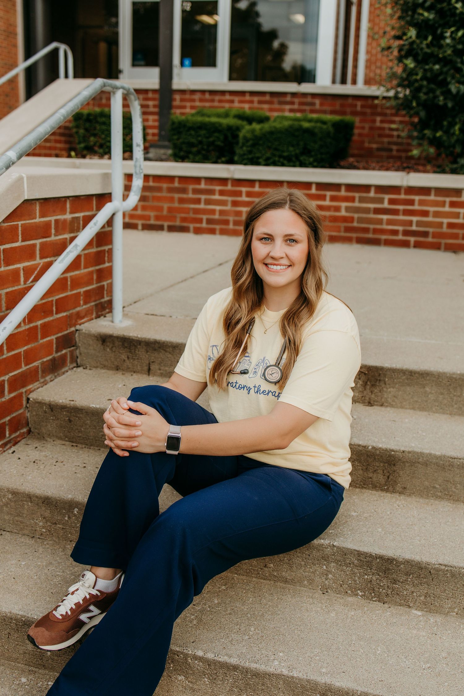 Young woman smiling on brick building steps, wearing yellow graphic tee, navy pants, and brown New Balance sneakers.