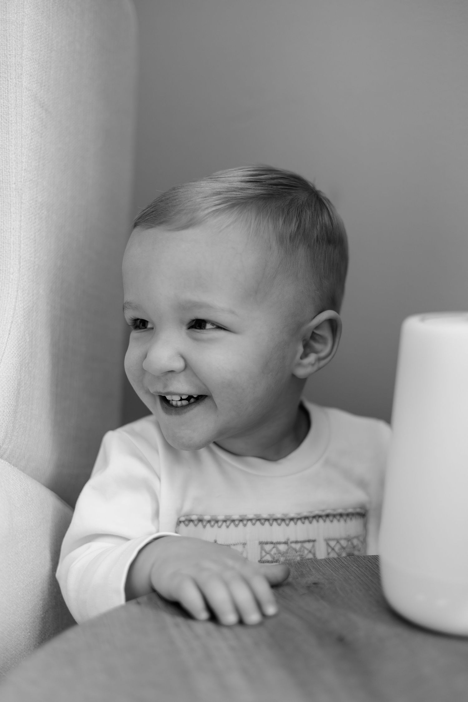 Black and white photo of a happy laughing toddler boy sitting at a wooden table next to a smart speaker.