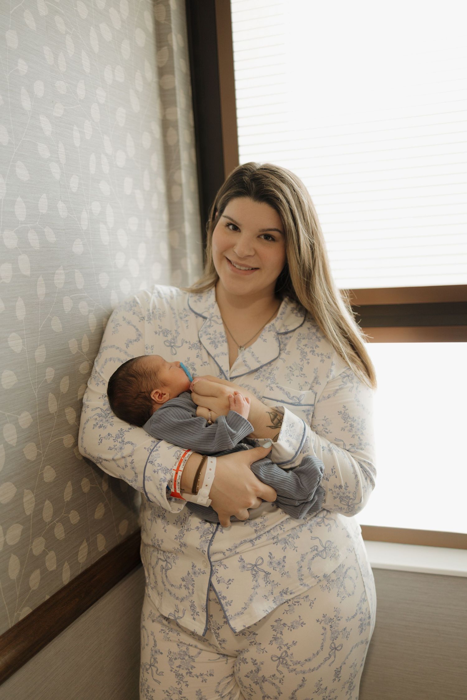 Smiling new mother in floral pajamas holds her newborn baby while standing near a window in a hospital room.