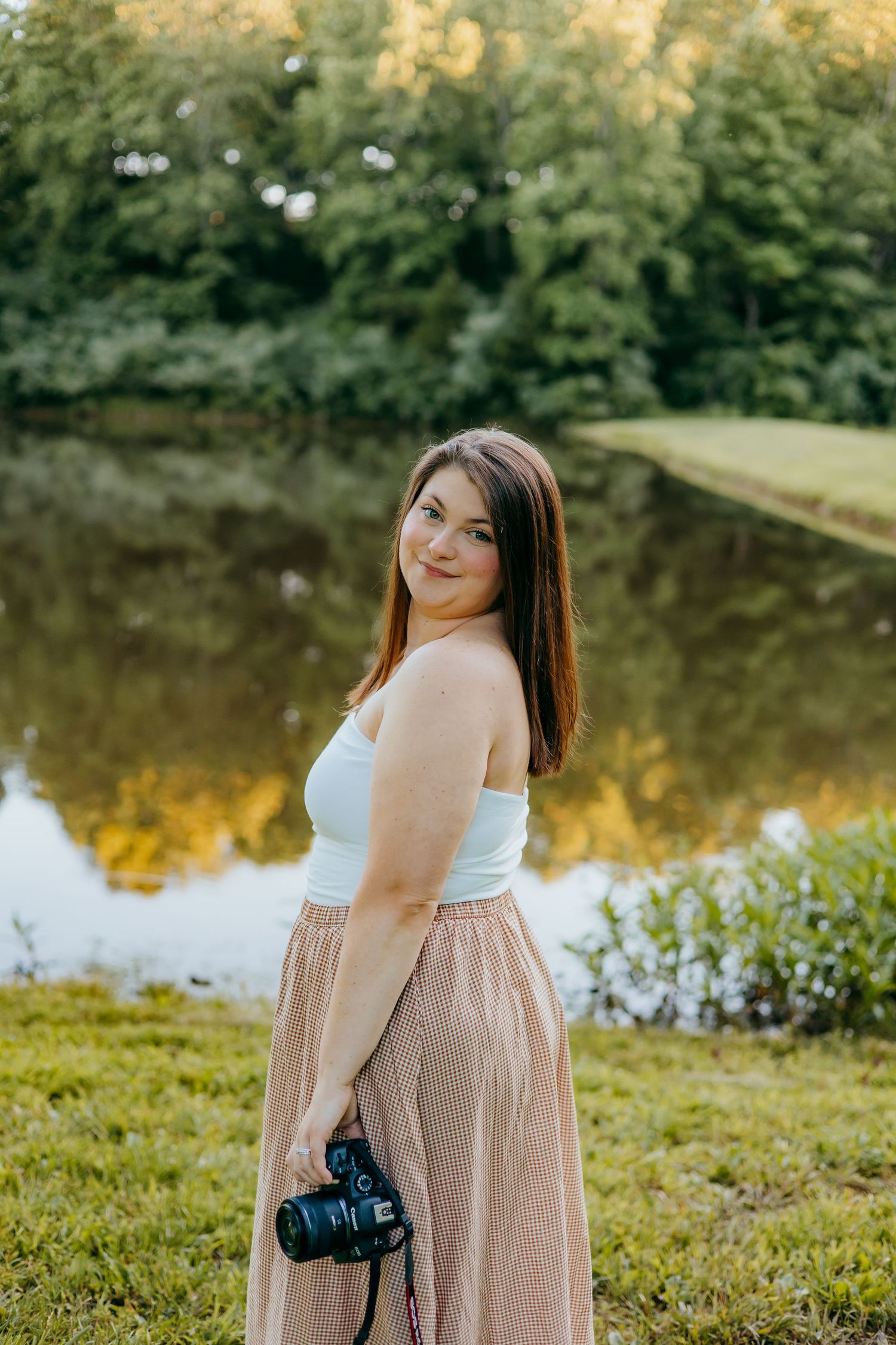 Woman in white top and plaid skirt holding a camera, smiling outdoors near a reflective pond with lush green trees.
