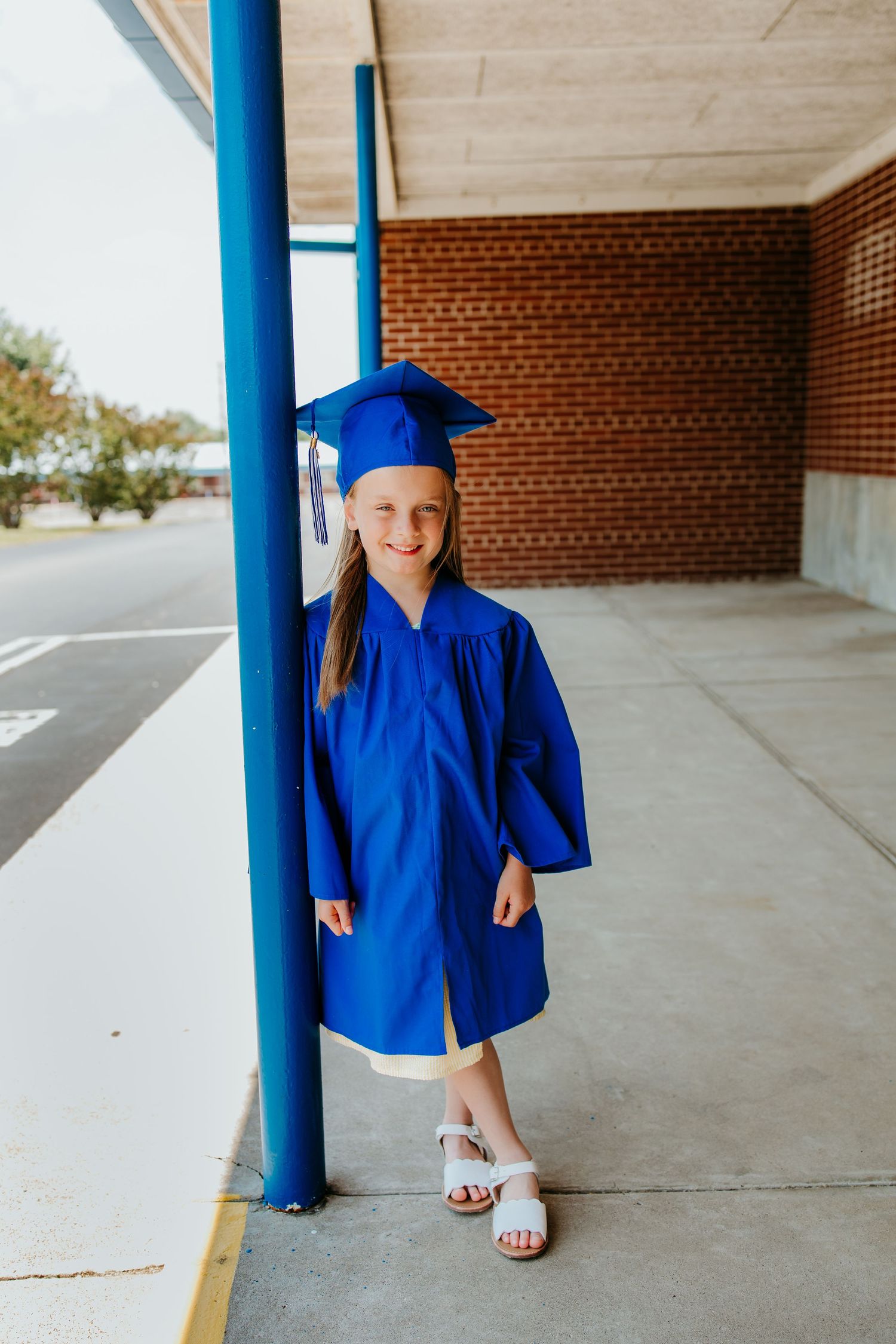 Young girl in blue graduation cap and gown smiling beside a blue pole outside a brick school building.