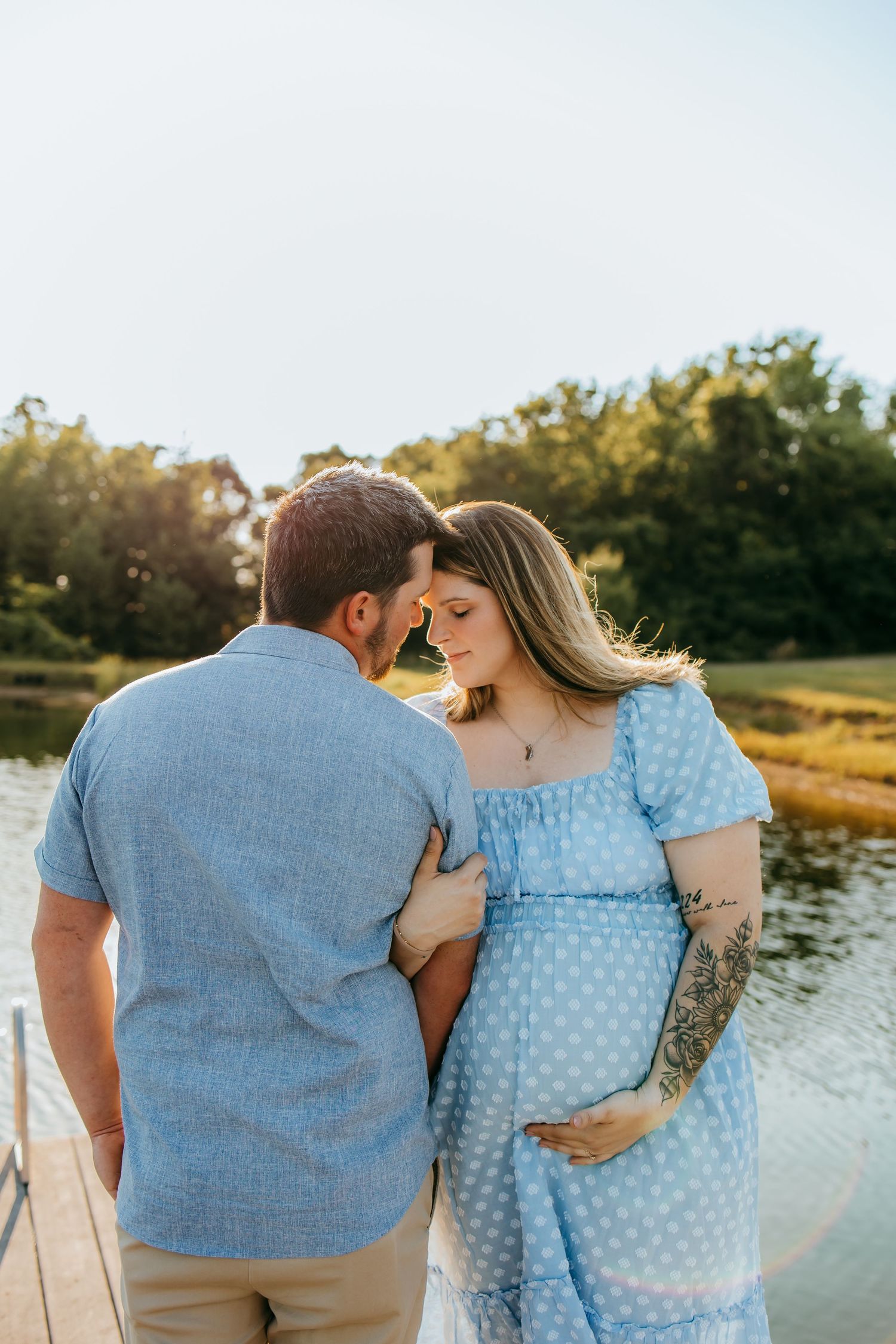 Pregnant couple sharing a tender moment on a dock by a lake at golden hour, woman in blue polka dot dress.