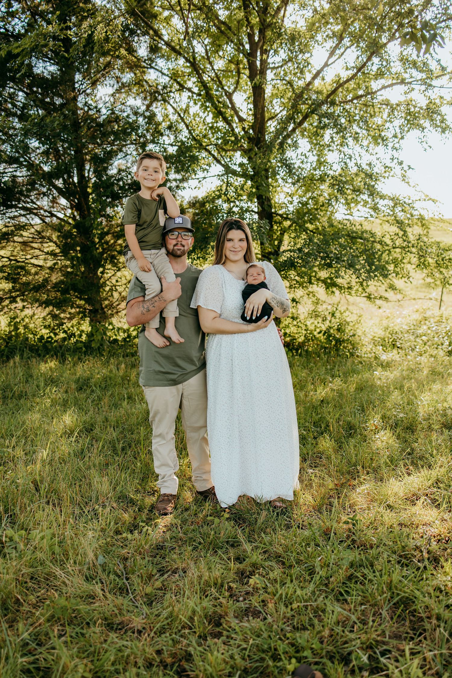 Family of four poses outdoors in a sunny green field, dad holding toddler on shoulders, mom cradling newborn in white dress.
