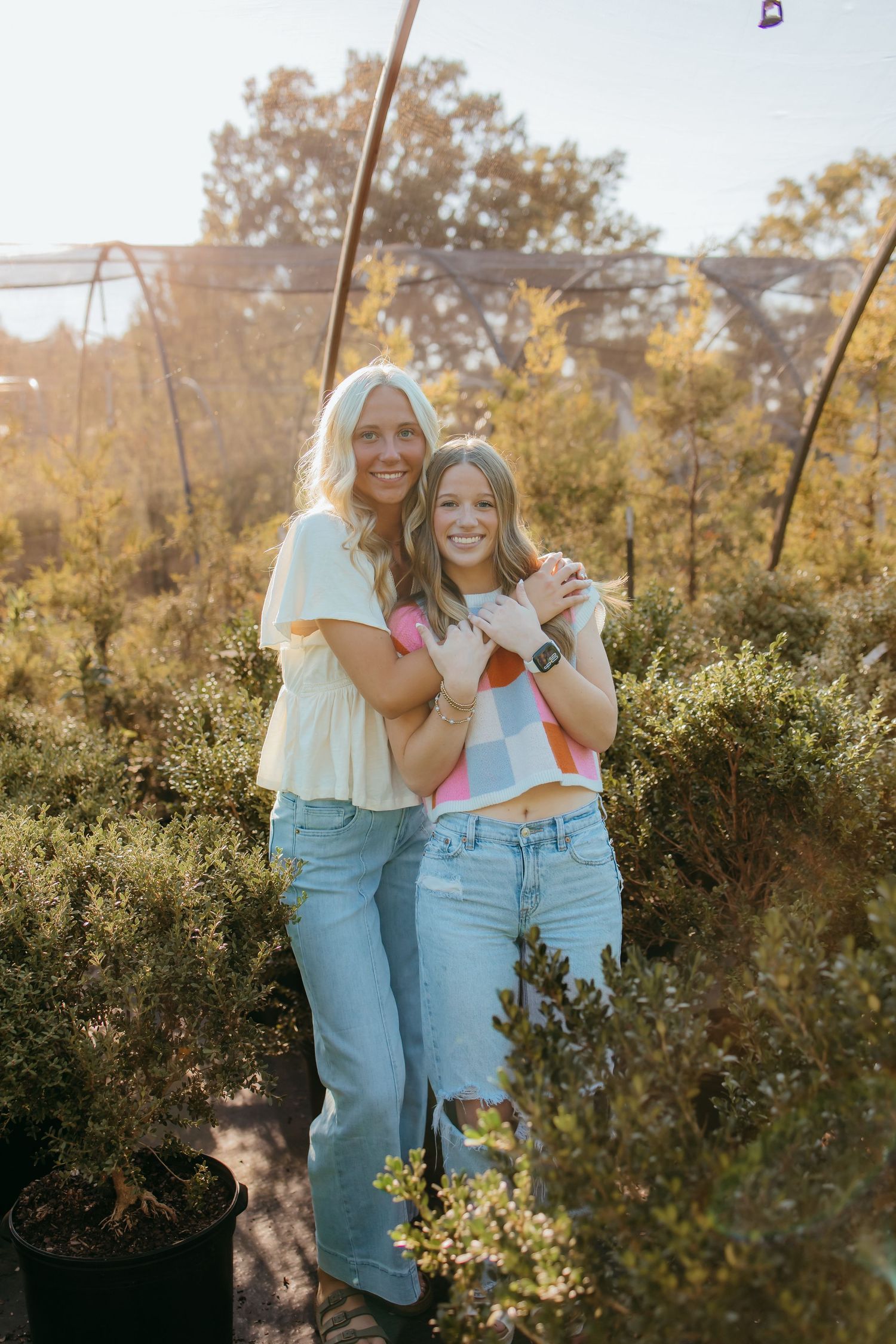 Two seniors in jeans pose together among green shrubs in golden hour sunlight outdoors.