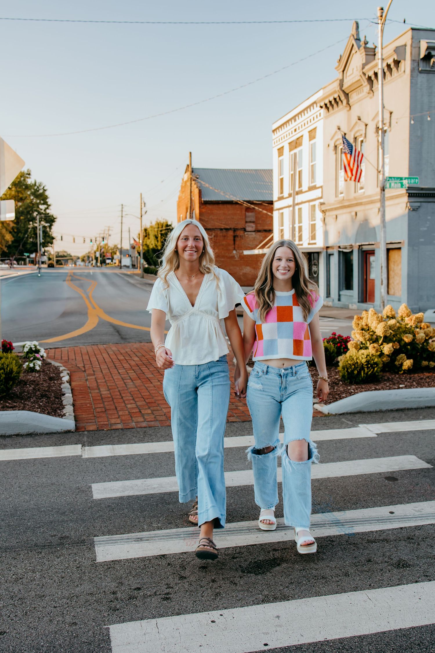 Two seniors in jeans cross a sunny small-town street crosswalk, one in white blouse, one in colorful crop top.