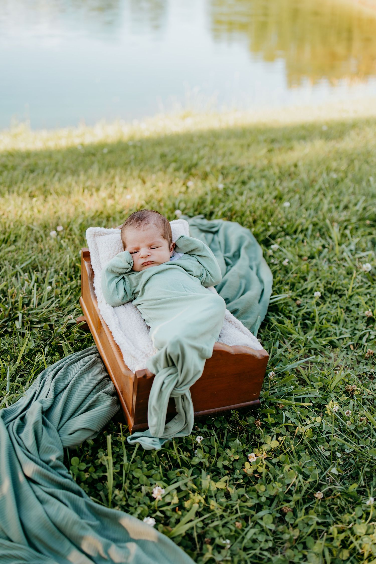 Newborn baby sleeping peacefully in a wooden crib outdoors on green grass near a lake, wrapped in a soft sage green blanket.