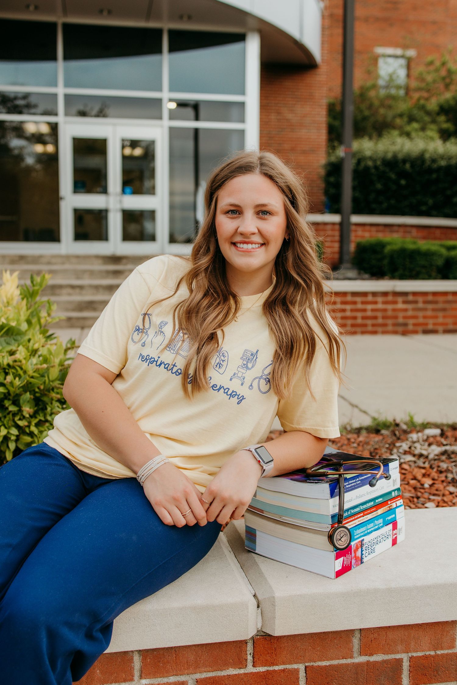 Student in respiratory therapy shirt sits smiling on brick ledge with textbooks outside university building.
