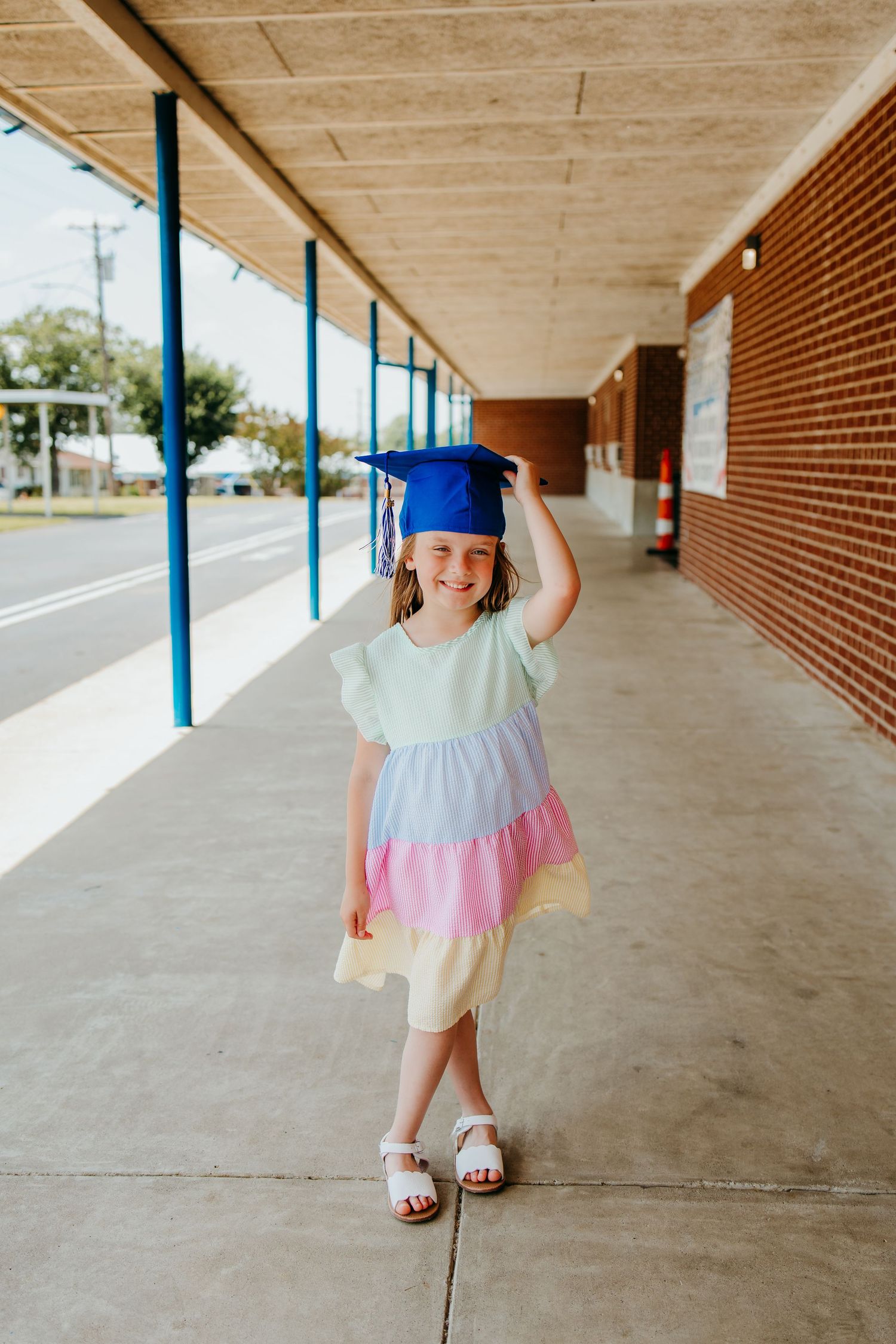 Young girl in colorful pastel dress and blue graduation cap smiling on school sidewalk.