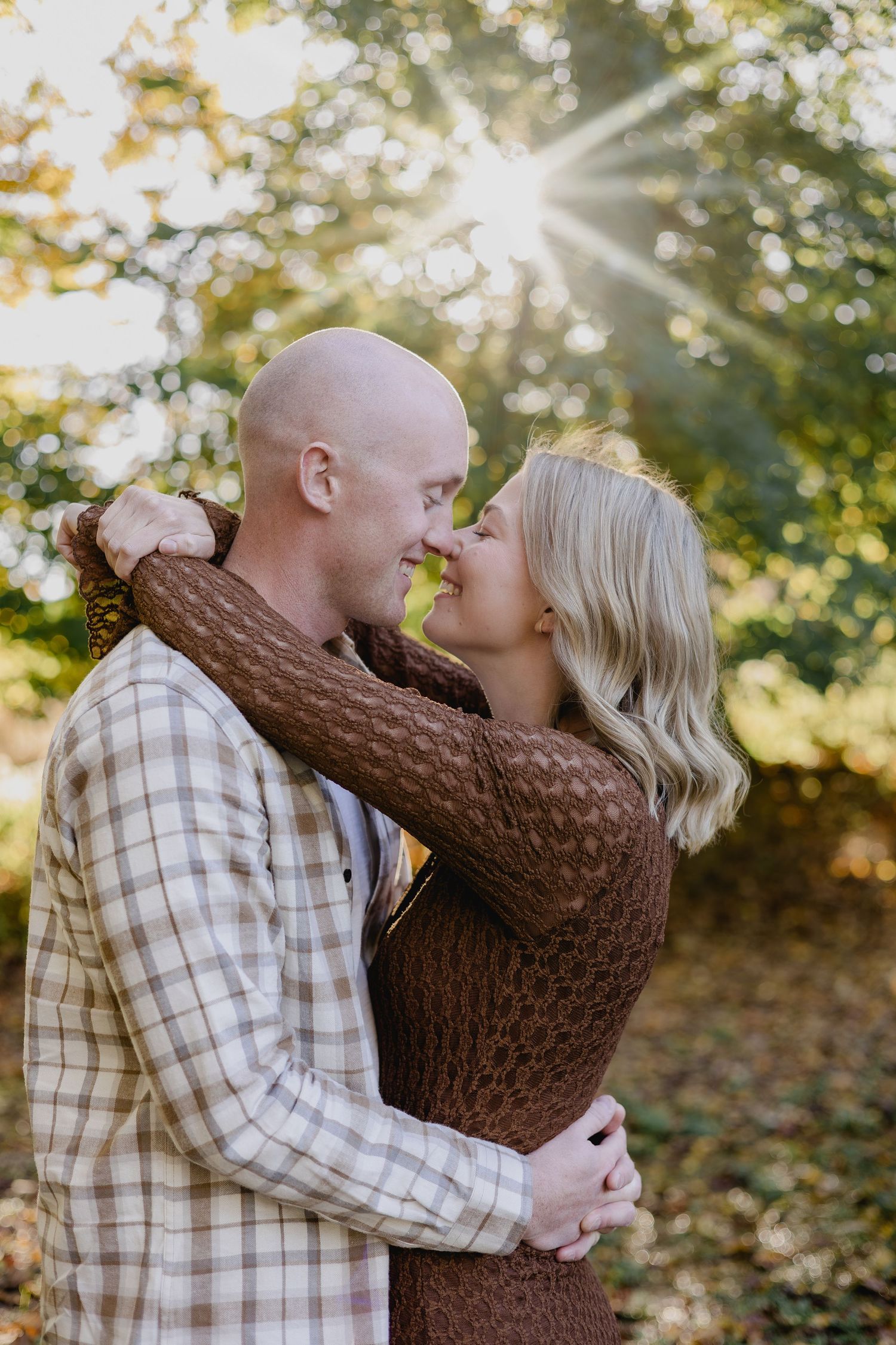 A couple embraces and nearly kisses outdoors in golden autumn light, surrounded by lush green trees.