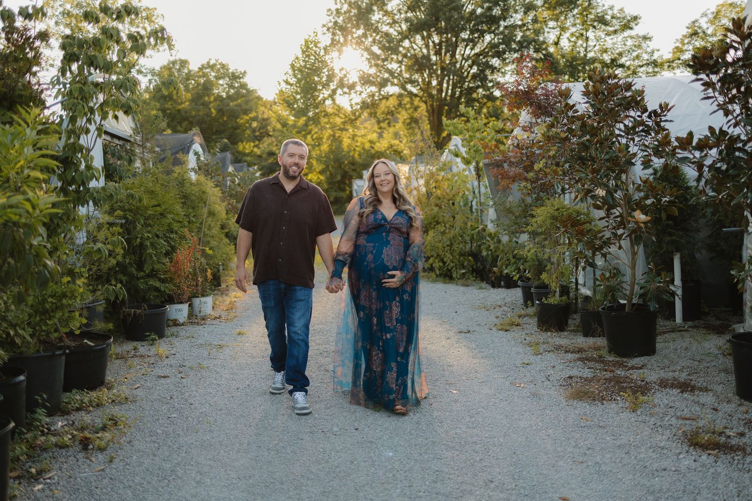 Pregnant couple walking hand-in-hand on a gravel path surrounded by lush greenery and potted trees in golden hour light.